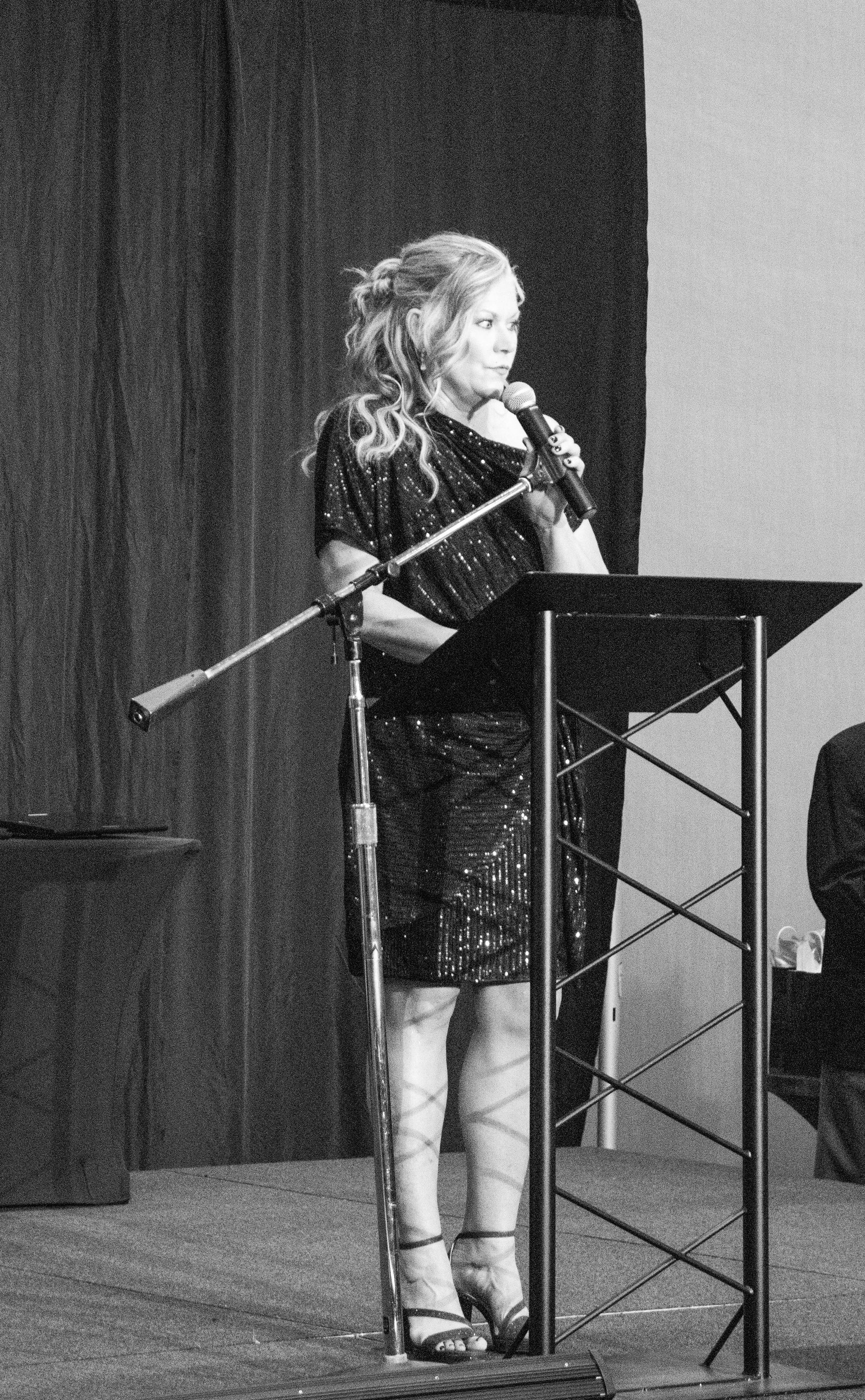 A person with wavy hair standing at a lectern, speaking into a microphone on a stage with a dark curtain background.