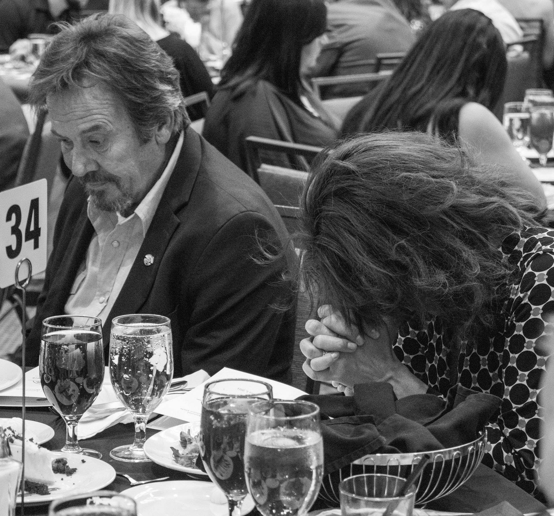 A man looks down at a table while a person beside him sits with head bowed and hands clasped in prayer at a formal dinner.