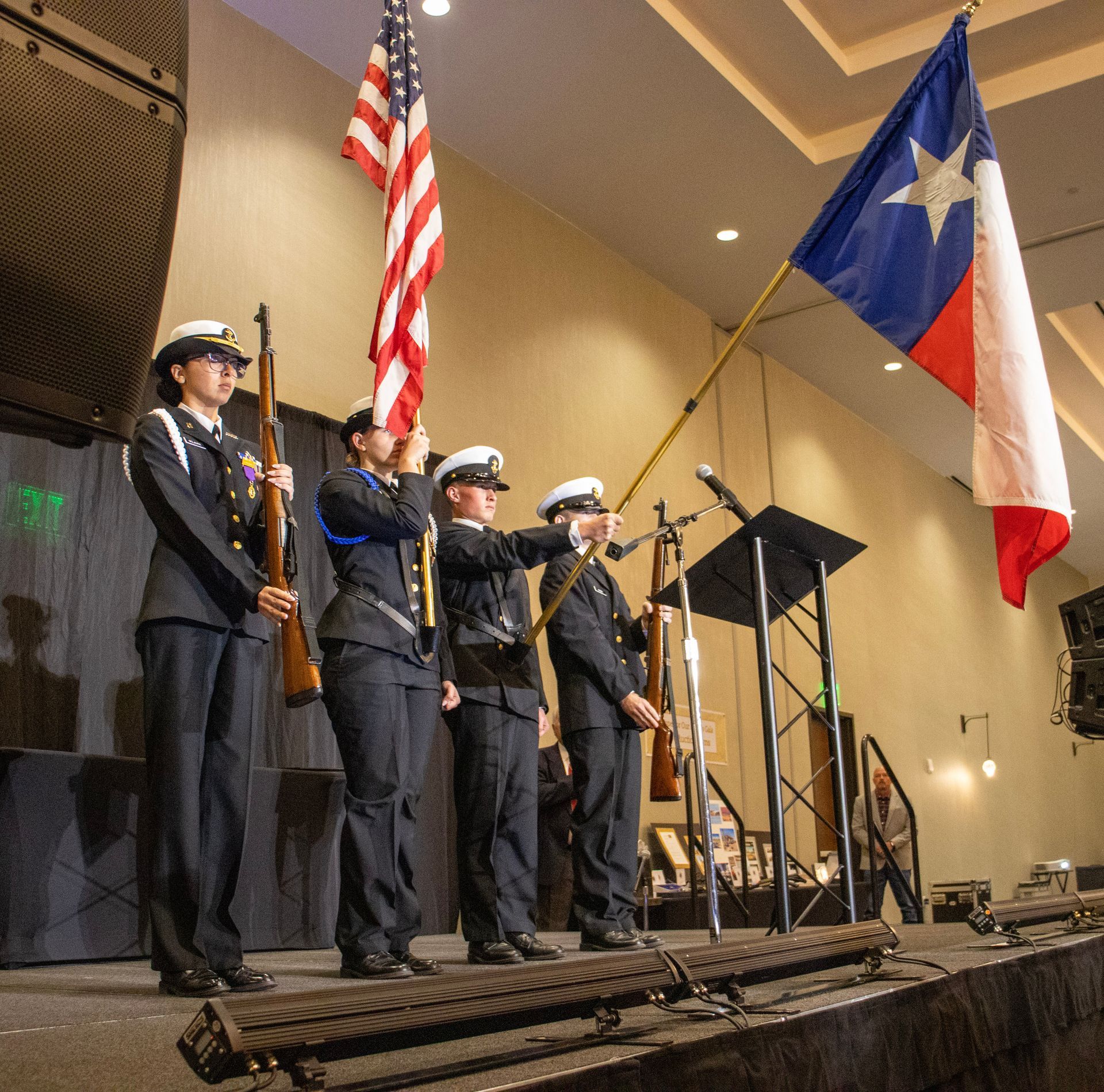 Four individuals in military-style uniforms perform a color guard ceremony with US and Texas flags on a stage.