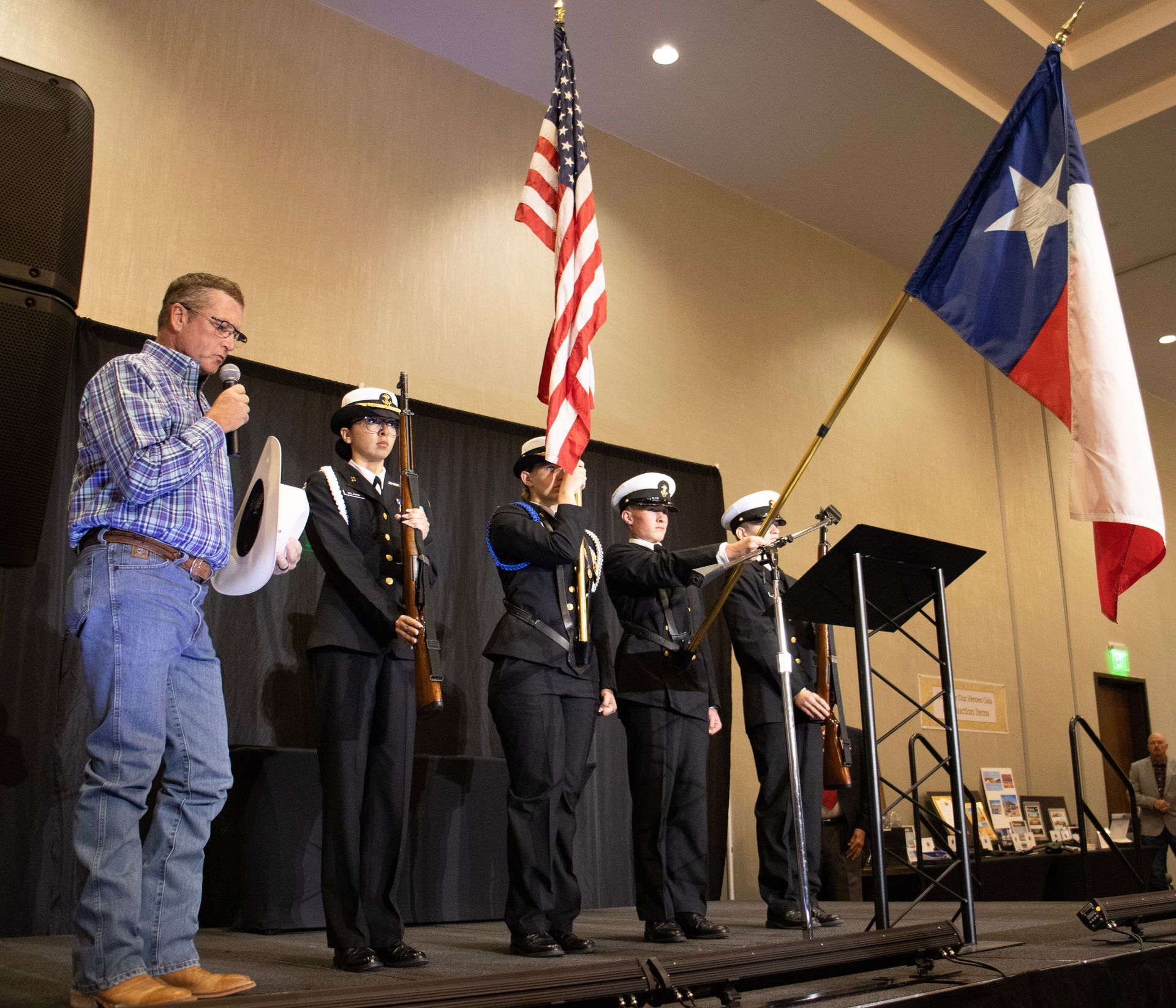 A speaker stands next to a four-person color guard holding American and Texas flags on a stage during an event.