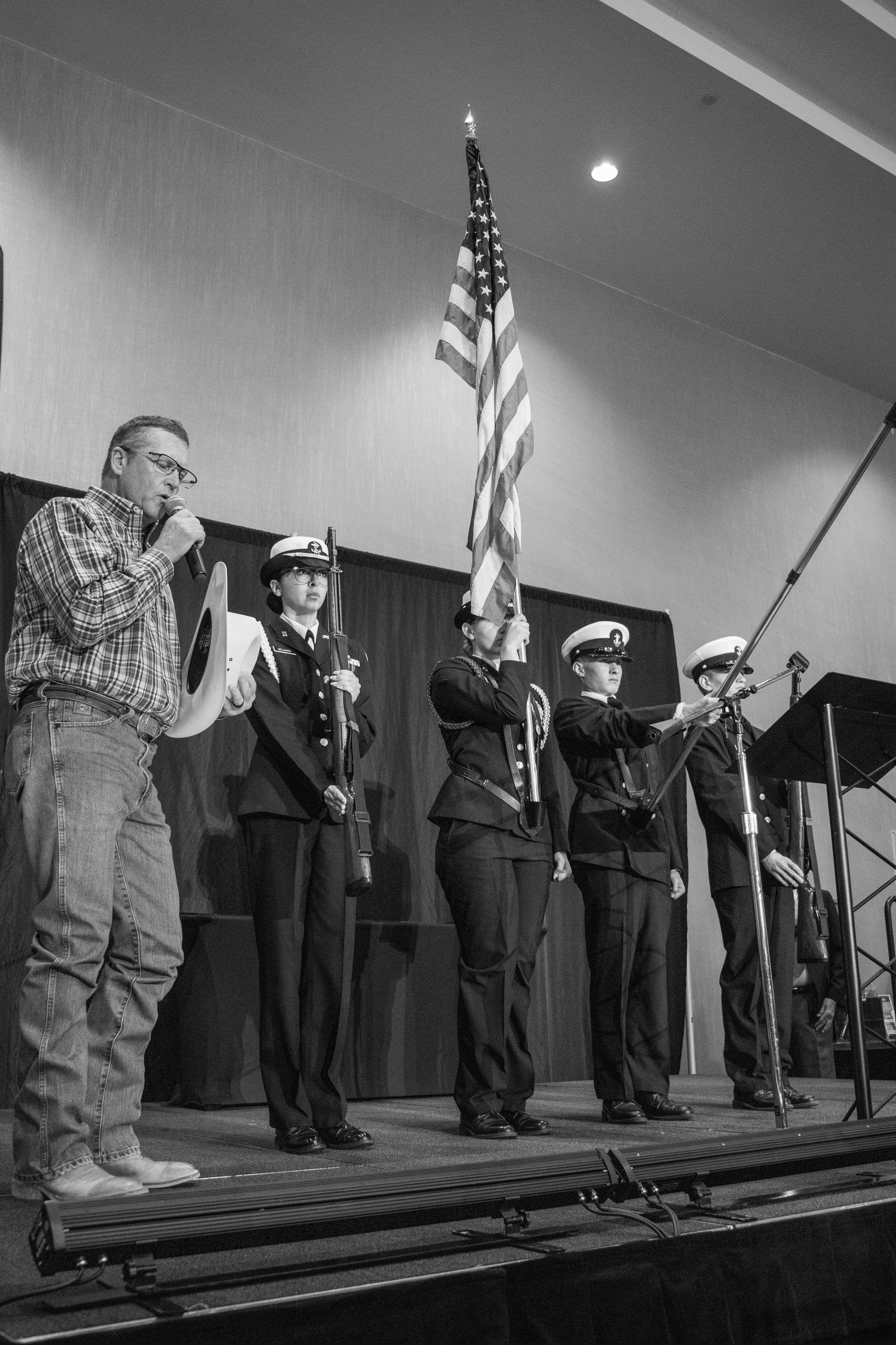 A man speaks at a microphone while a color guard stands behind him on stage, holding the American flag.
