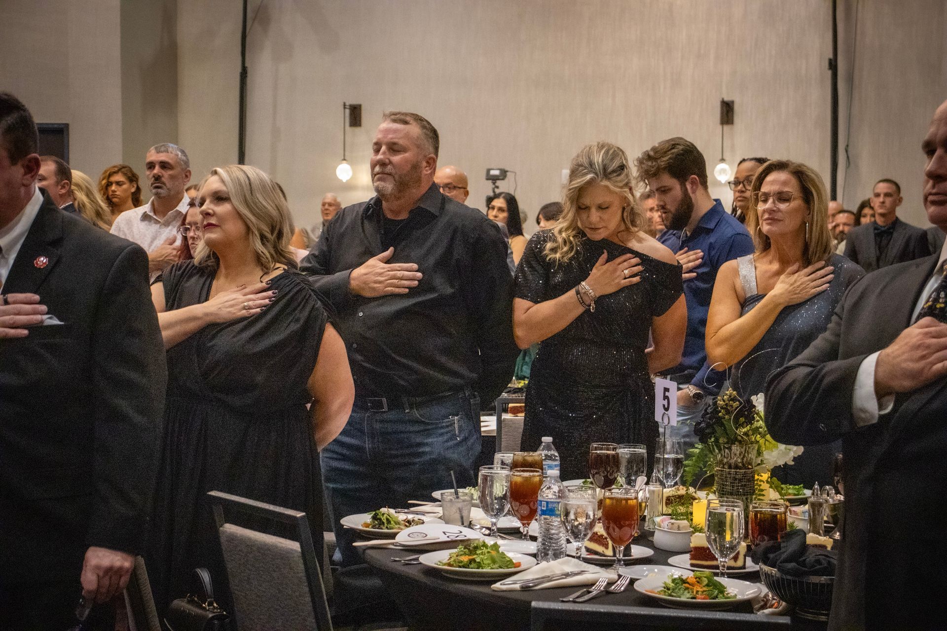 A group of people at a formal event standing with hands over their hearts, facing forward in a solemn posture.
