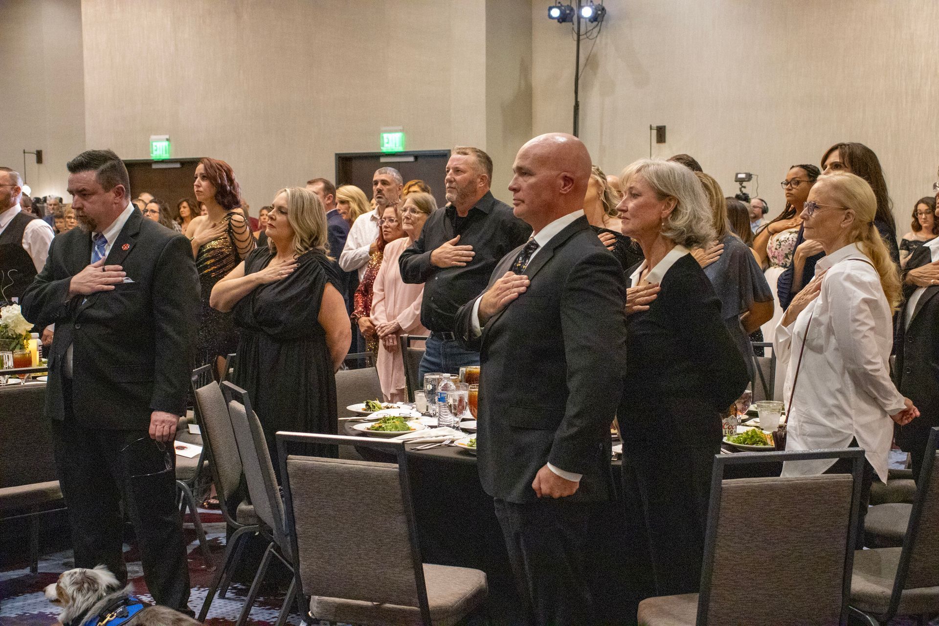 A group of people stands in a banquet hall with hands over their hearts, appearing to recite the Pledge of Allegiance.