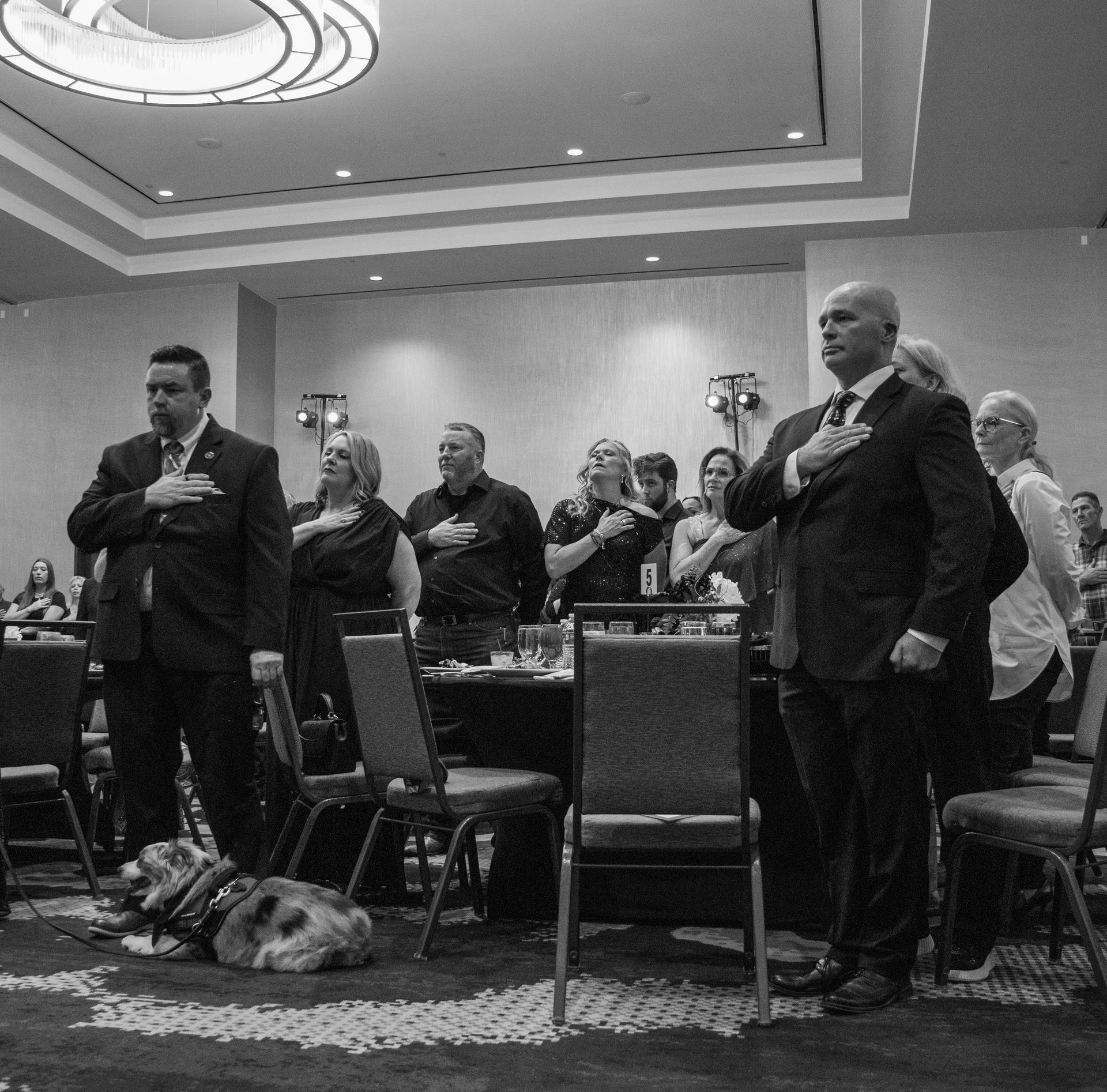 A group of people in formal attire stand with hands over their hearts during an event, with a service dog lying nearby.