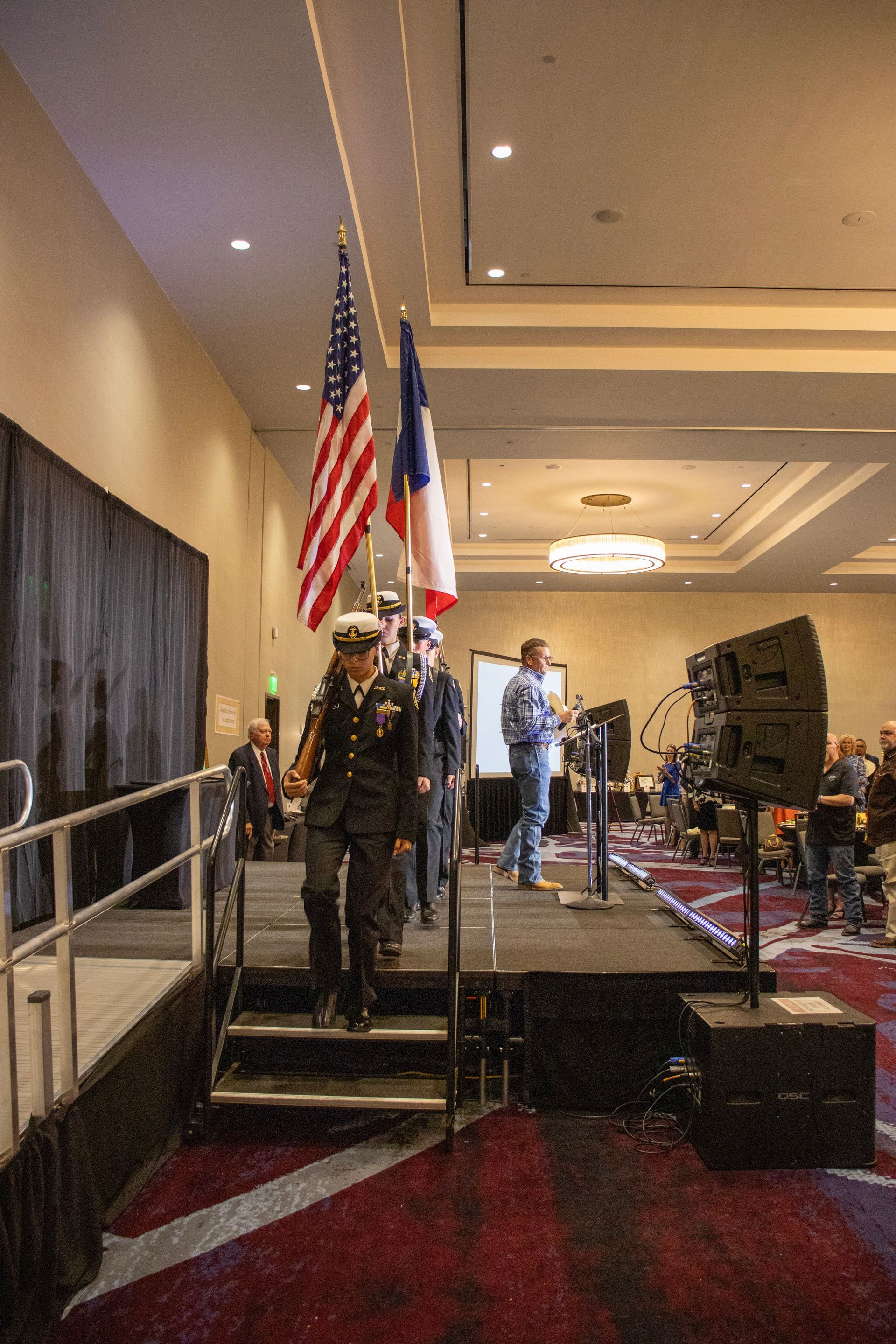 A color guard in uniform carrying the U.S. and Texas flags descends stairs on a stage at an indoor event.