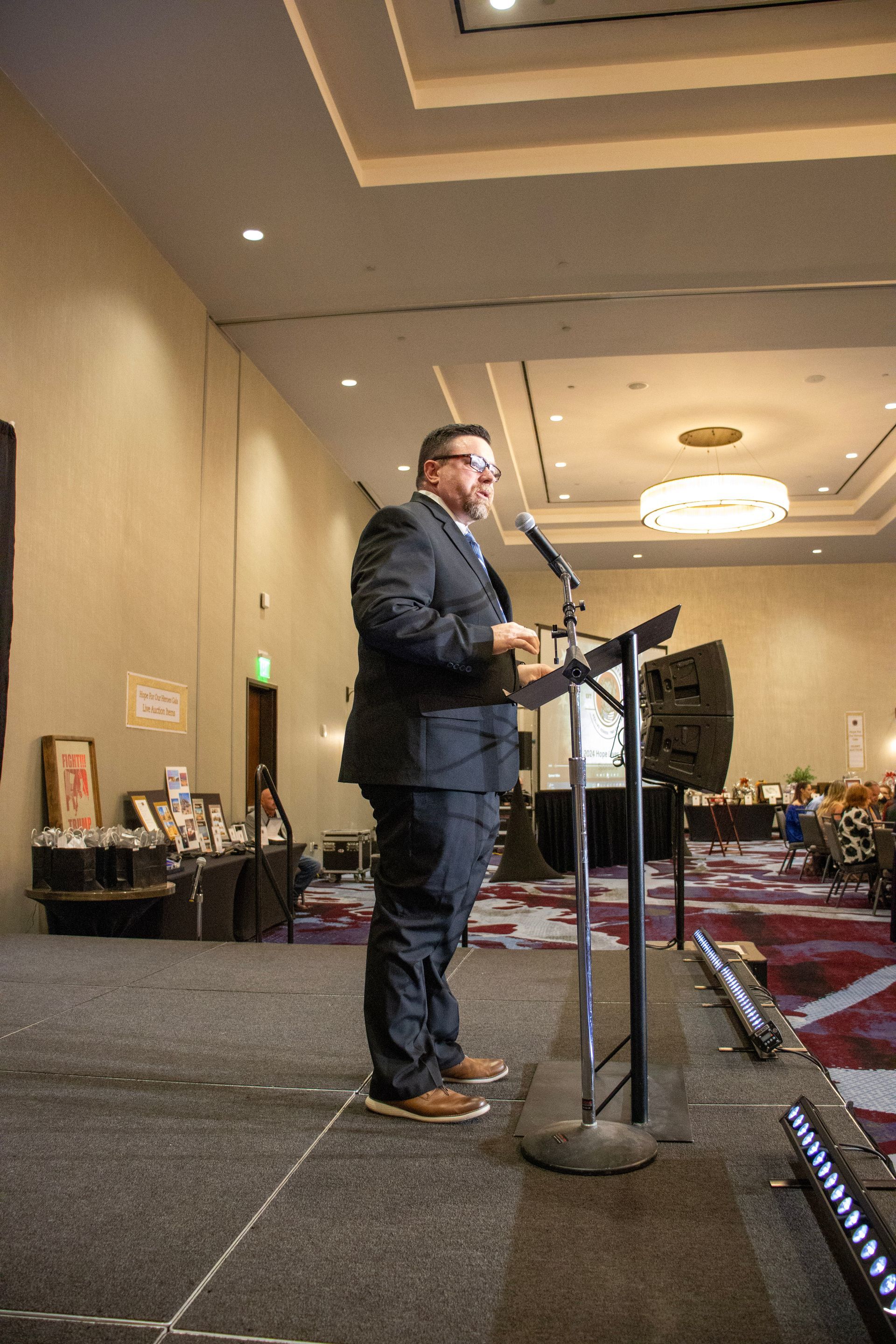 A person in a dark suit standing at a lectern on a stage, speaking into a microphone in a large, carpeted banquet hall.