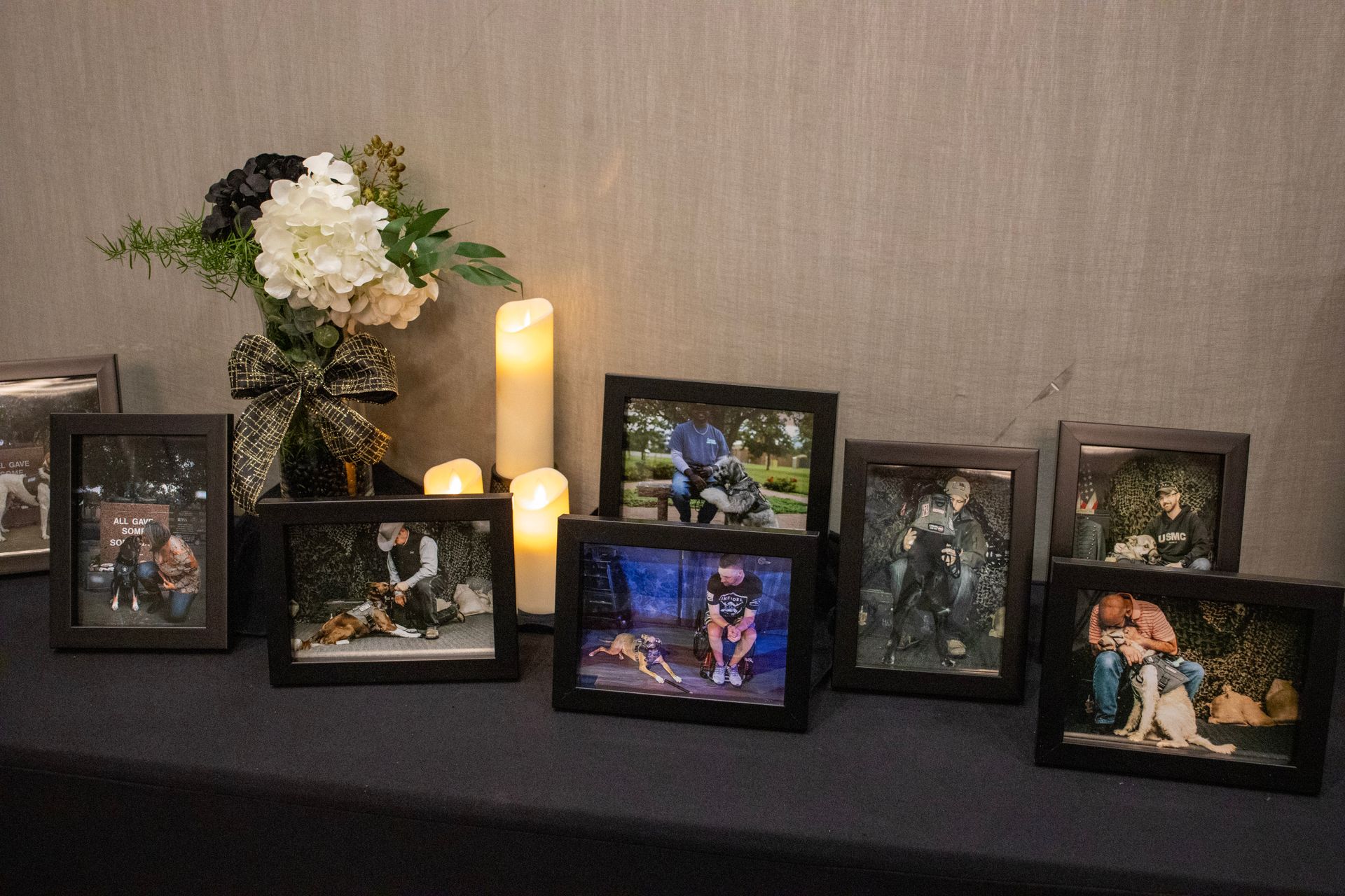 A memorial display featuring framed photos of a person with a dog, white flowers, and two candles on a black tablecloth.
