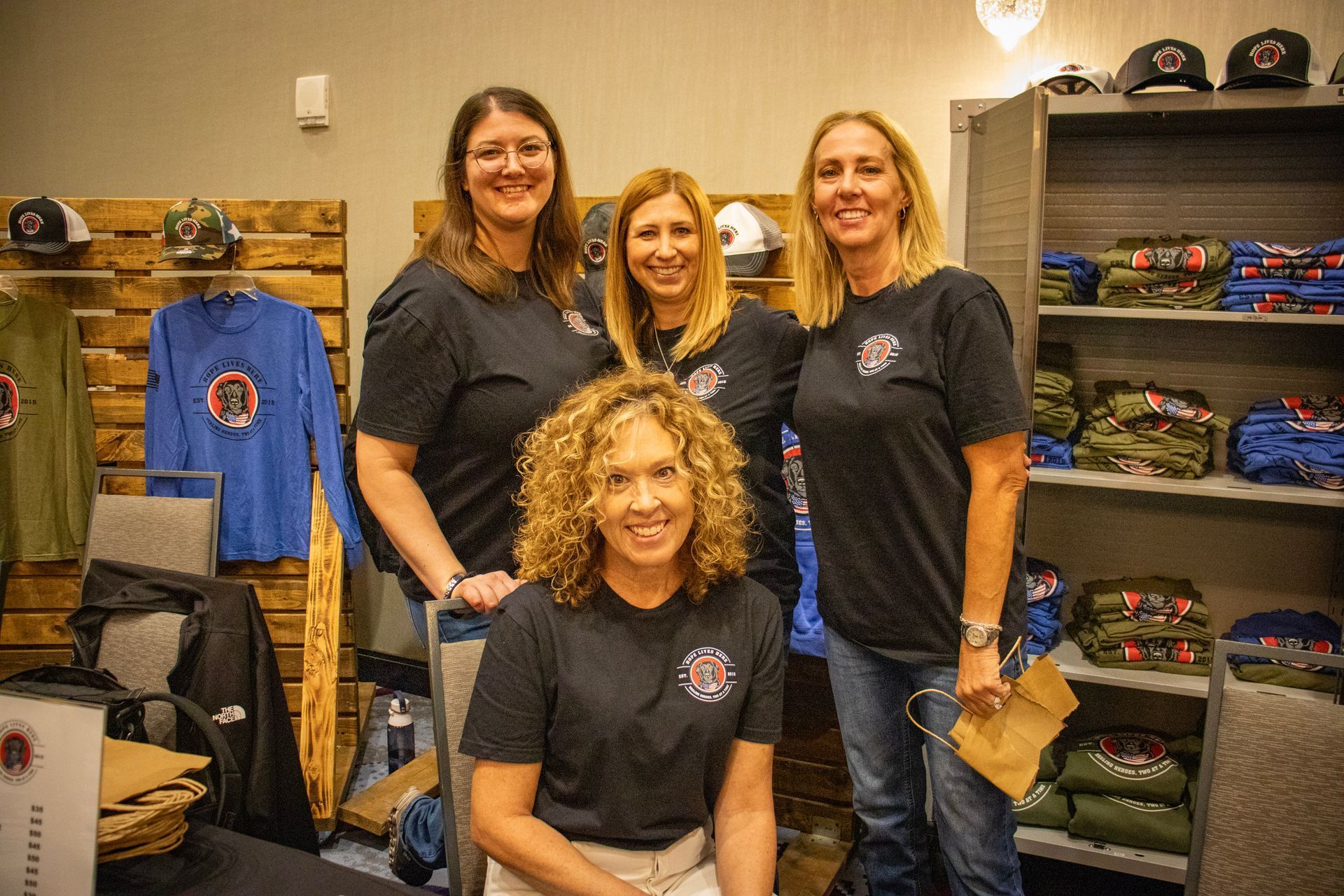 Four women in matching black logo t-shirts stand and sit in a retail space displaying hats and apparel.