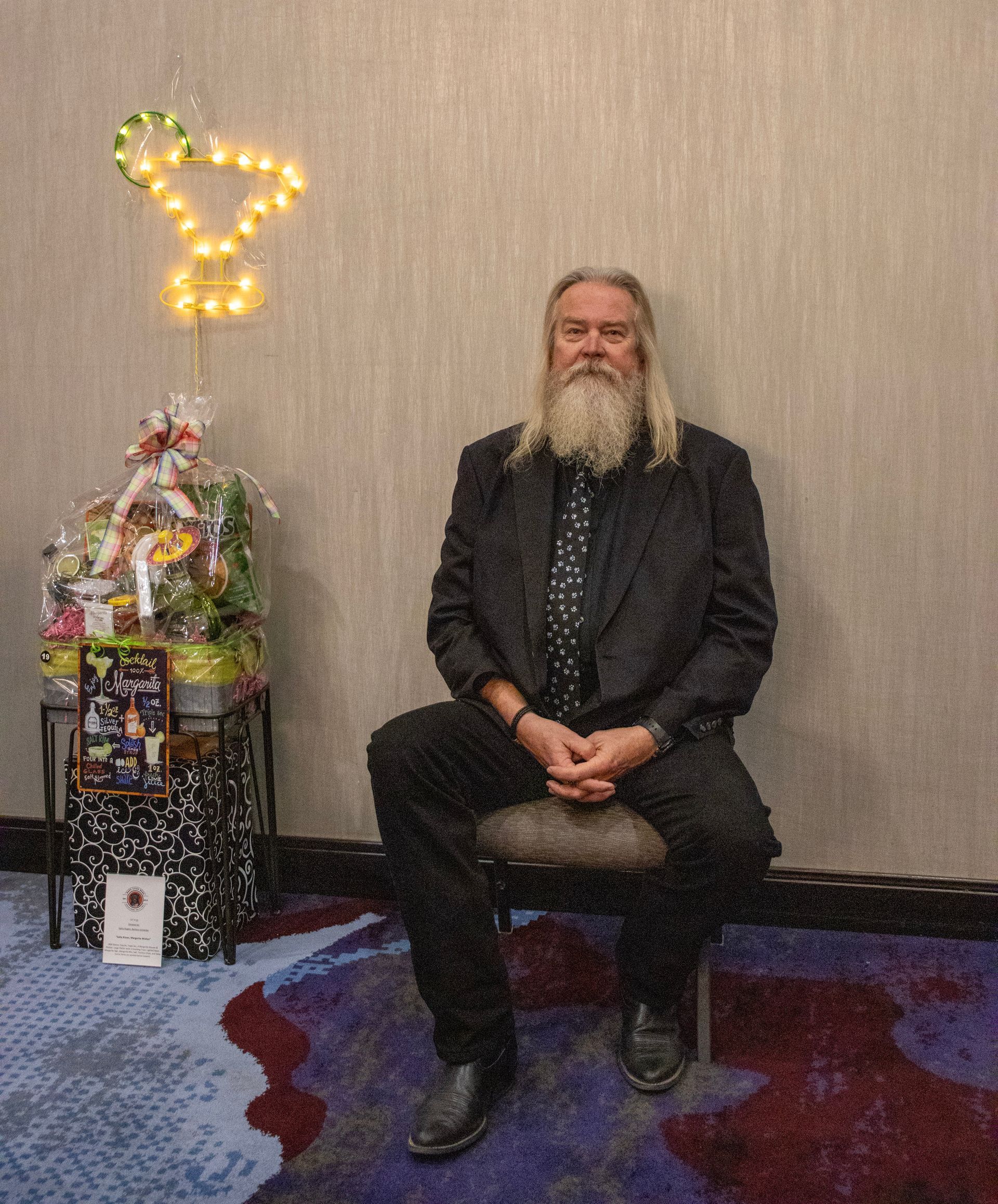 A long-bearded man in a dark suit sits on a chair next to a gift basket topped with a glowing martini glass decoration.