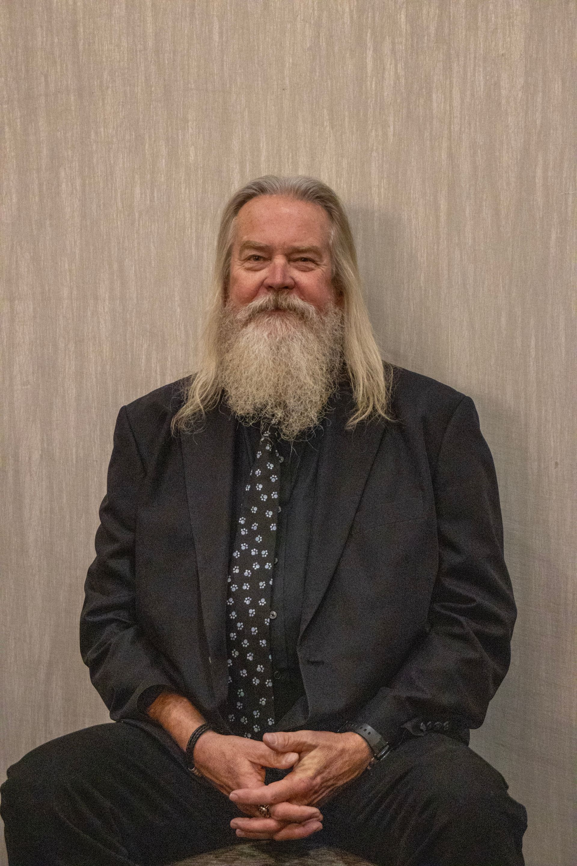 A person with long gray hair and a beard, wearing a dark blazer and a patterned tie, sits against a light textured wall.