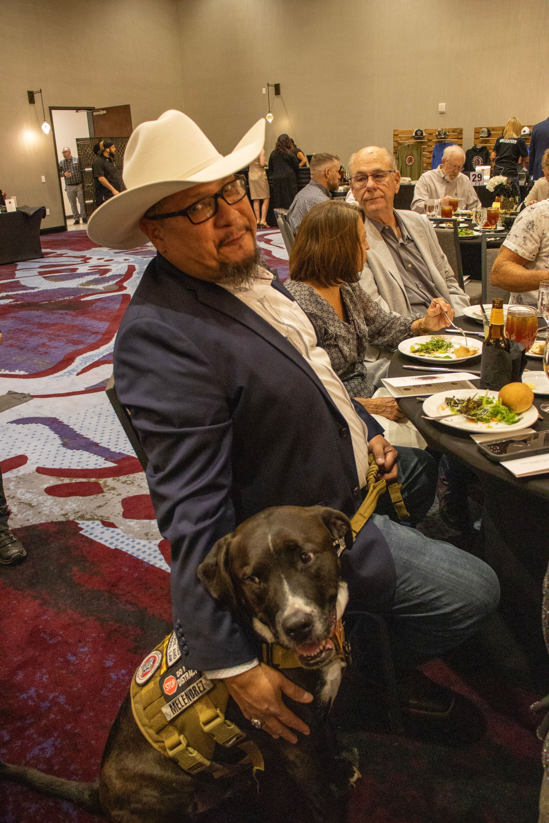 A man in a cowboy hat and blazer holds a service dog wearing a tactical vest at a table in a carpeted banquet hall.