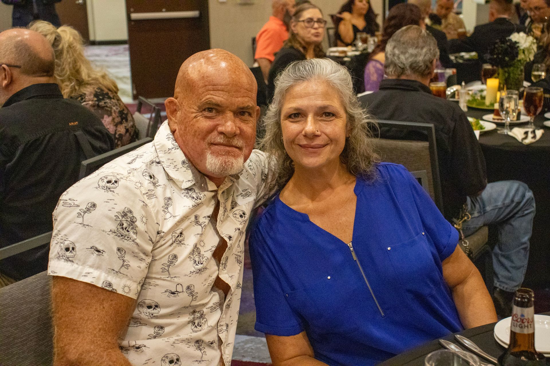 A smiling couple poses for a photo at a dinner event, with guests and tables visible in the blurred background.
