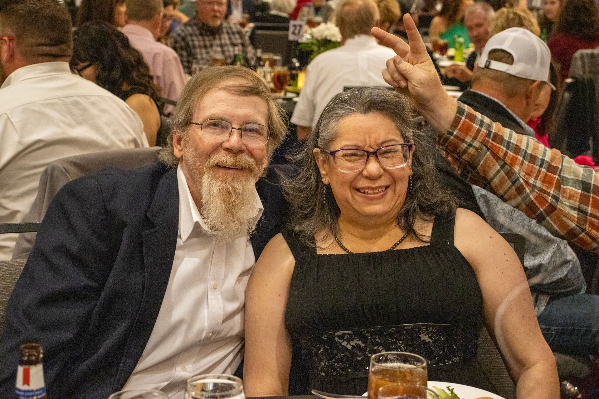 A smiling couple sits at a dinner table while someone behind them makes a peace sign gesture.