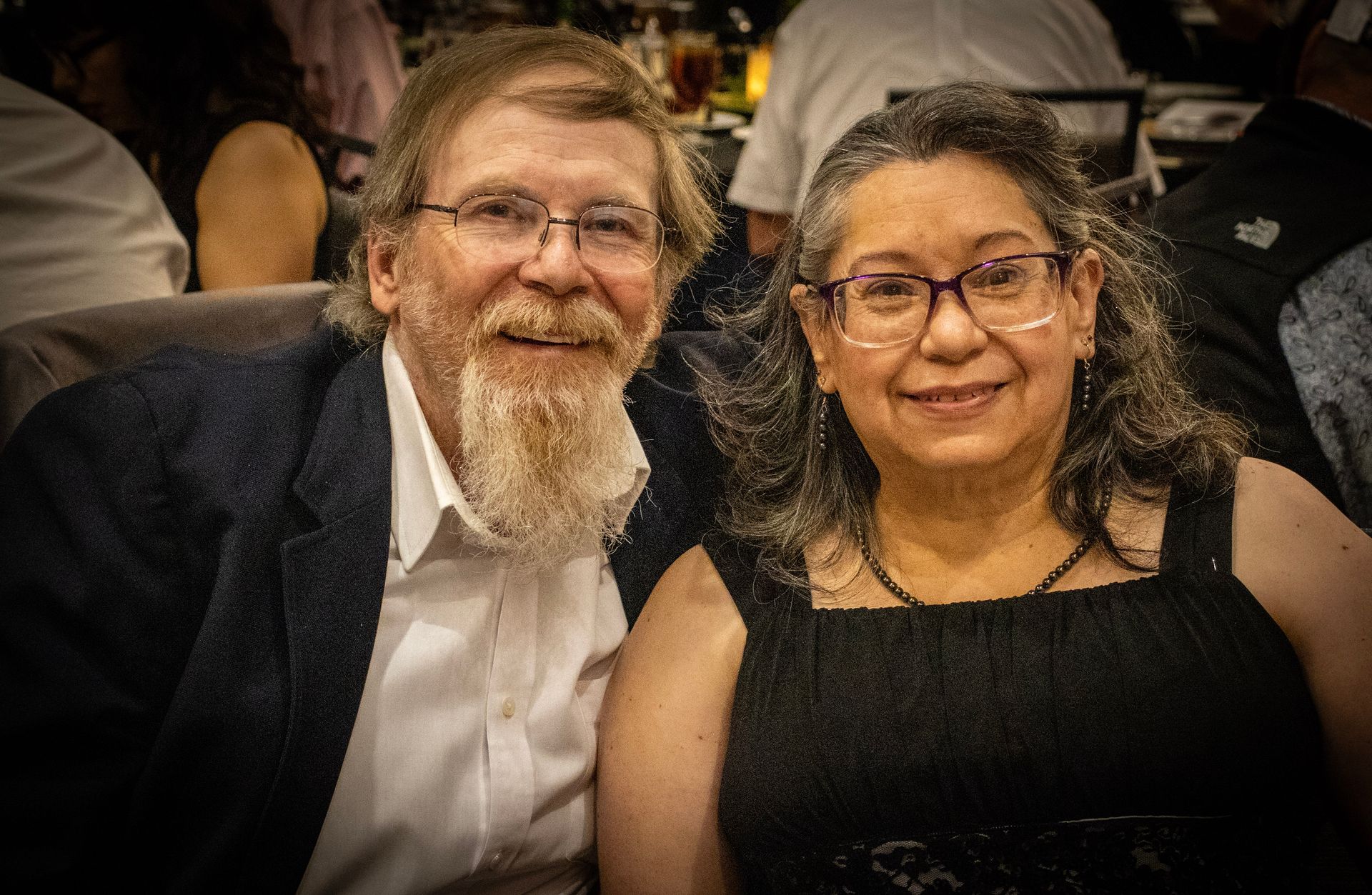 Two smiling people wearing glasses pose together at an indoor event, wearing a suit jacket and a black sleeveless dress.