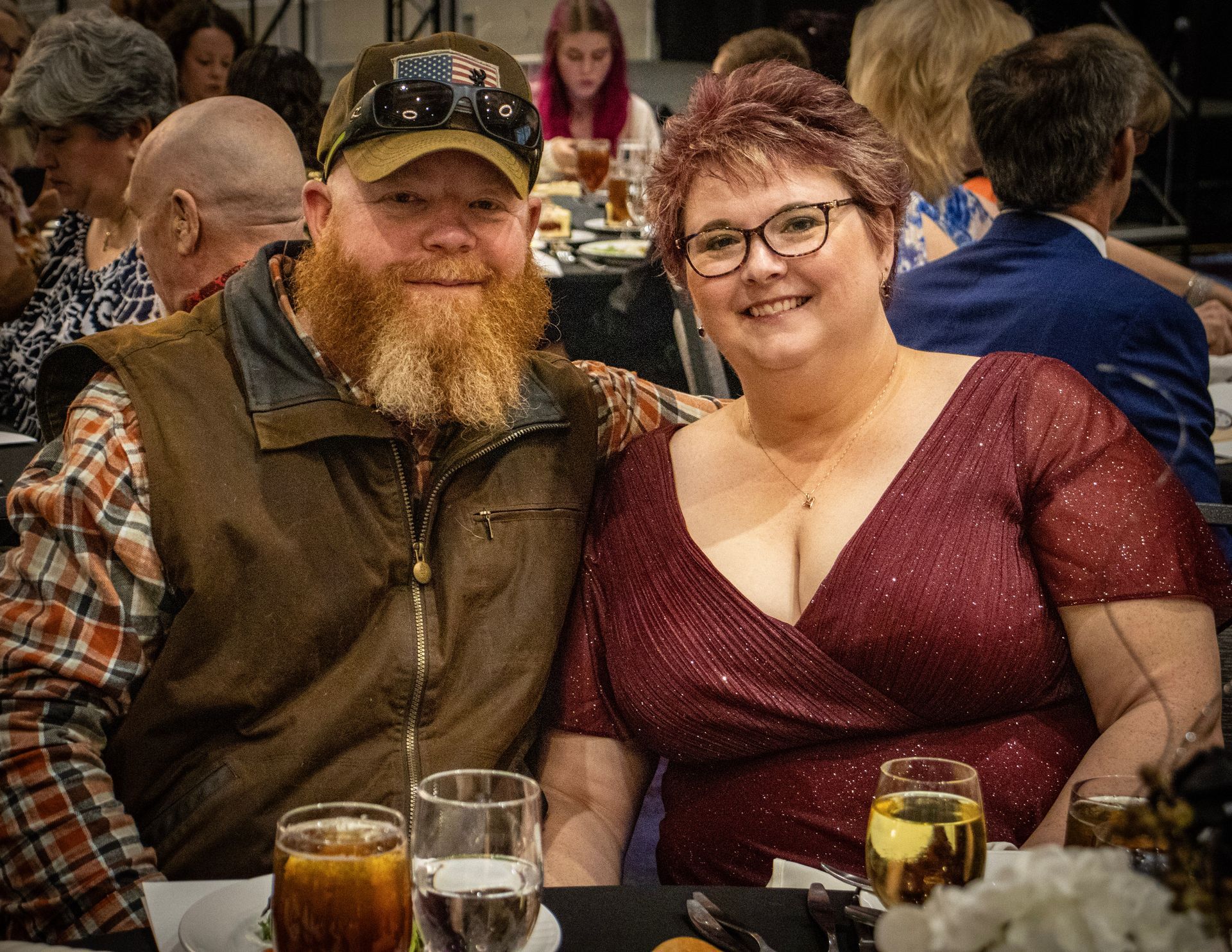 A smiling couple sits together at a table in a dimly lit, crowded venue. The man wears a cap and vest; the woman, glasses.