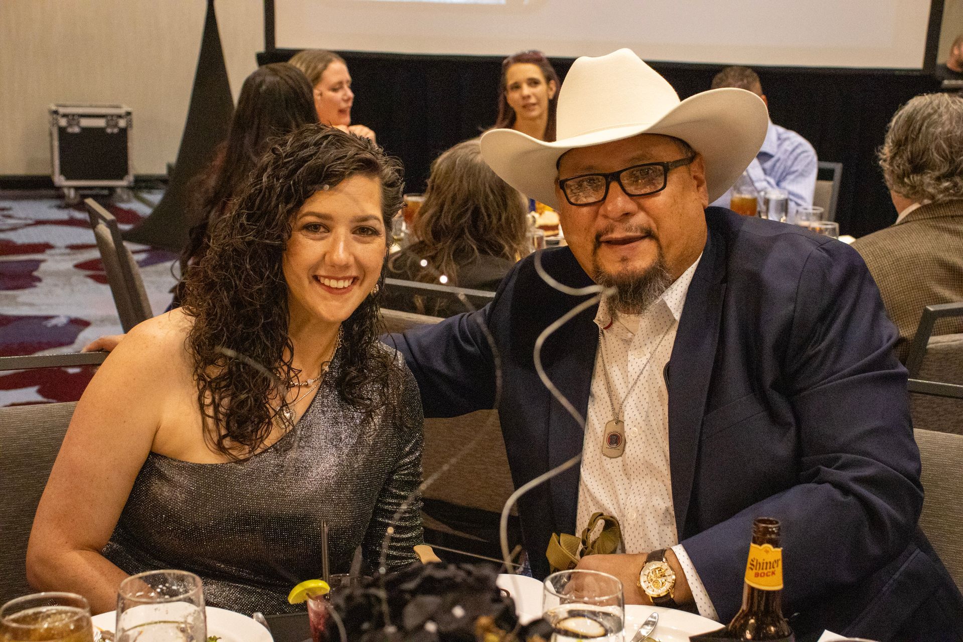 A smiling woman in a shimmery, one-shoulder top poses next to a man in a blazer and cowboy hat at a dinner event.