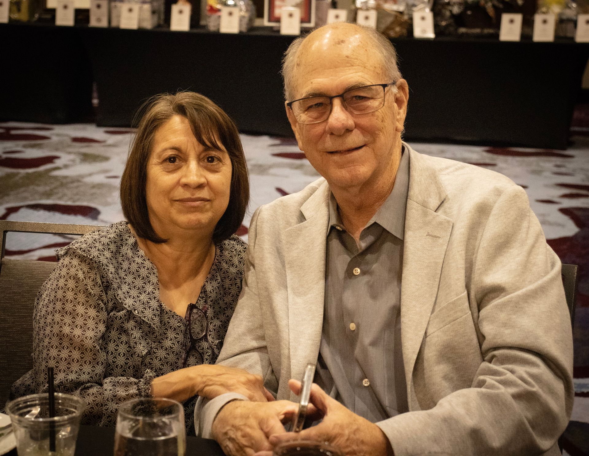A person with dark hair and a patterned blouse poses beside someone wearing glasses and a light blazer at a banquet.