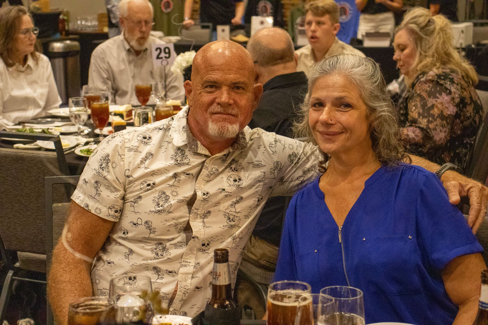 A couple smiling while sitting together at a table during a social event, surrounded by other attendees.