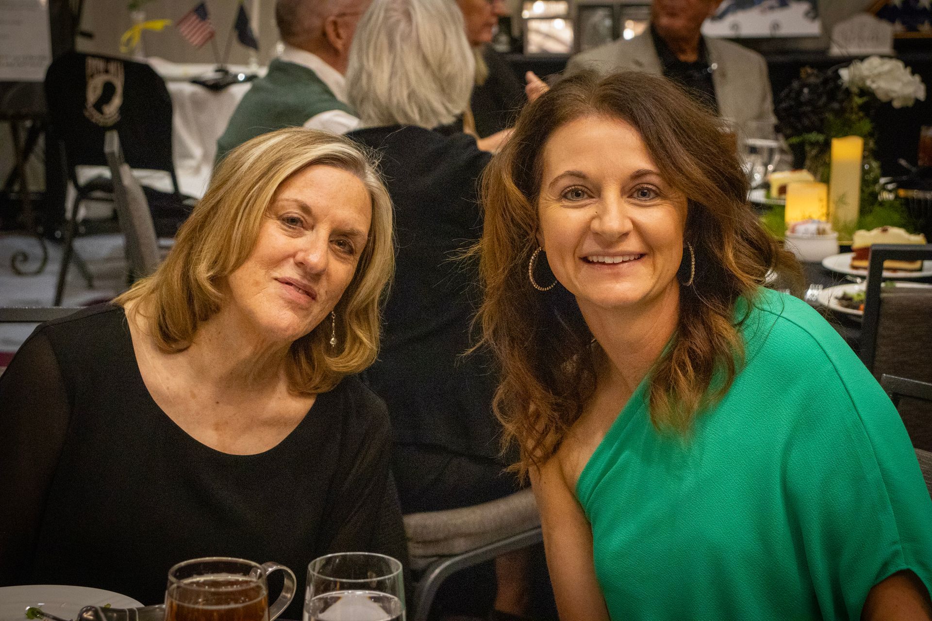 Two women smile at the camera while sitting at a table during an indoor event.