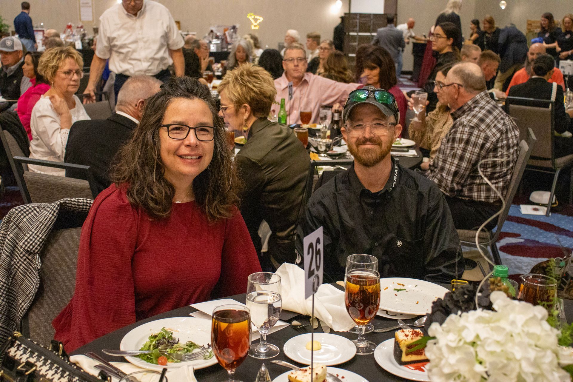 A person in a maroon sweater and a person in a black shirt sit at a table with a number 26 card at a banquet event.