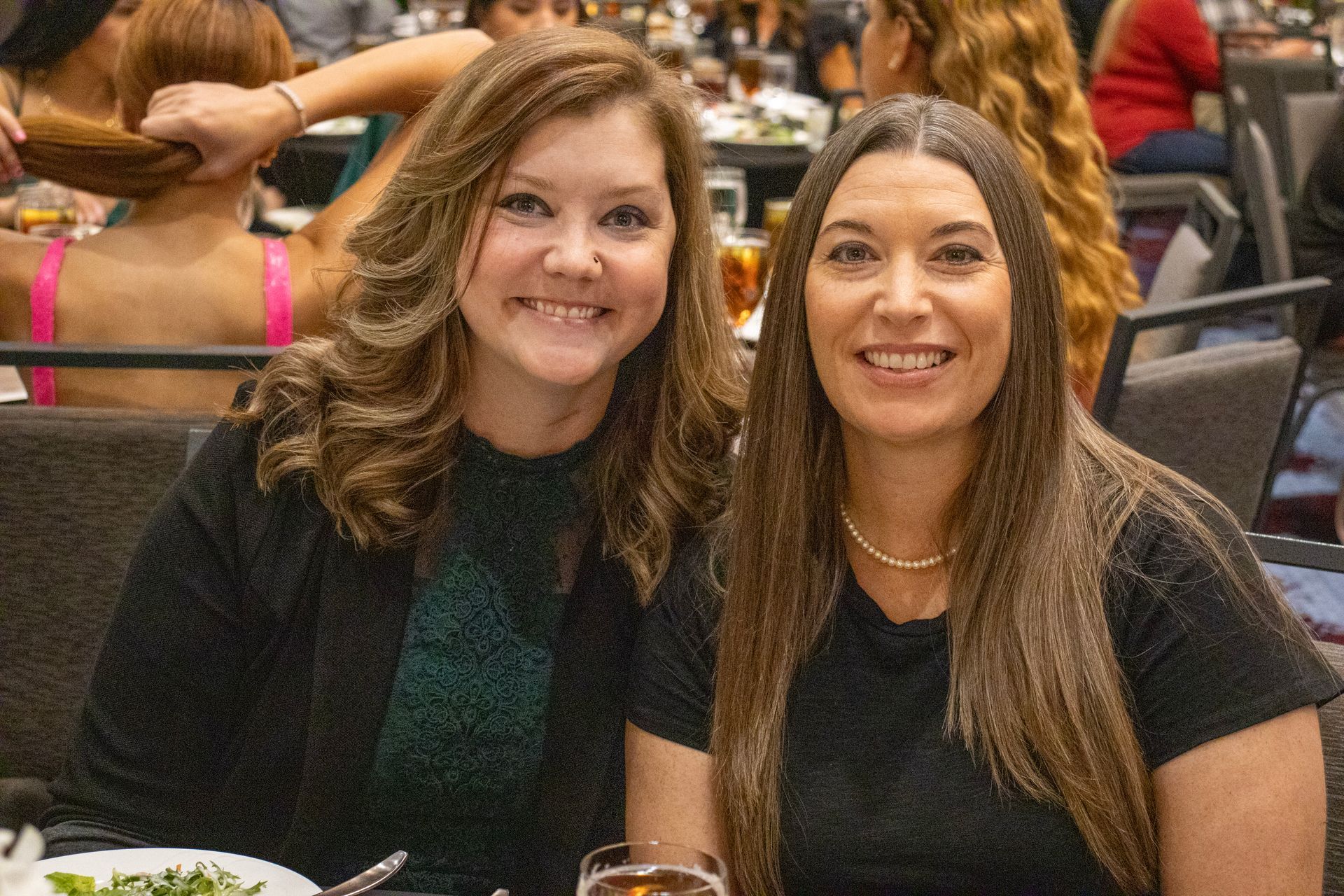 Two smiling individuals sit together at a table in a crowded banquet hall, both facing the camera.