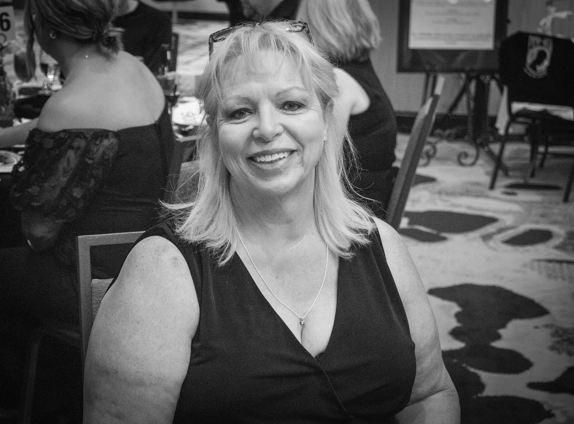 A smiling woman with blonde hair wearing a black sleeveless dress, seated at a formal event.