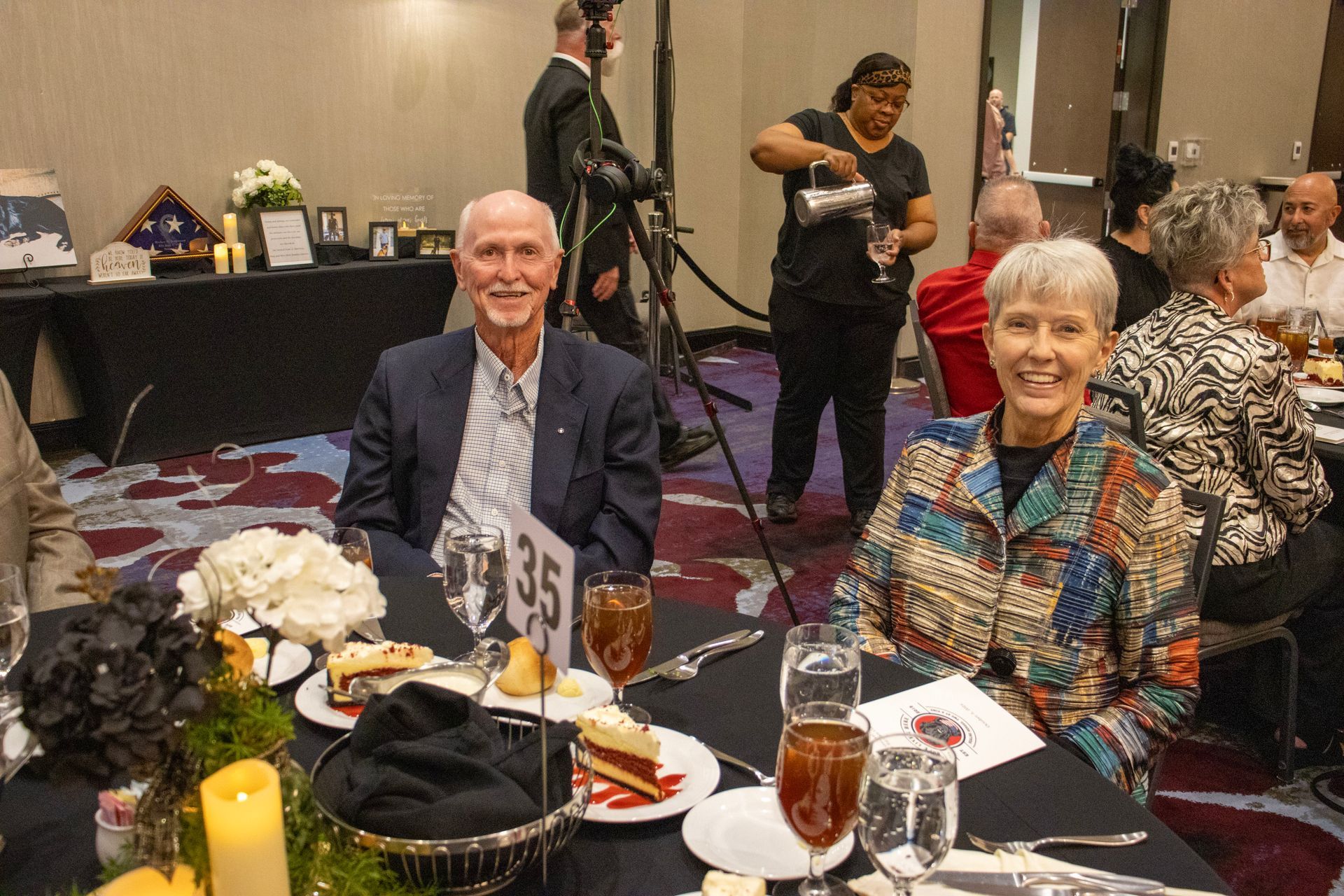 A man and woman smile at a banquet table while a server pours a drink in the background of a formal event.