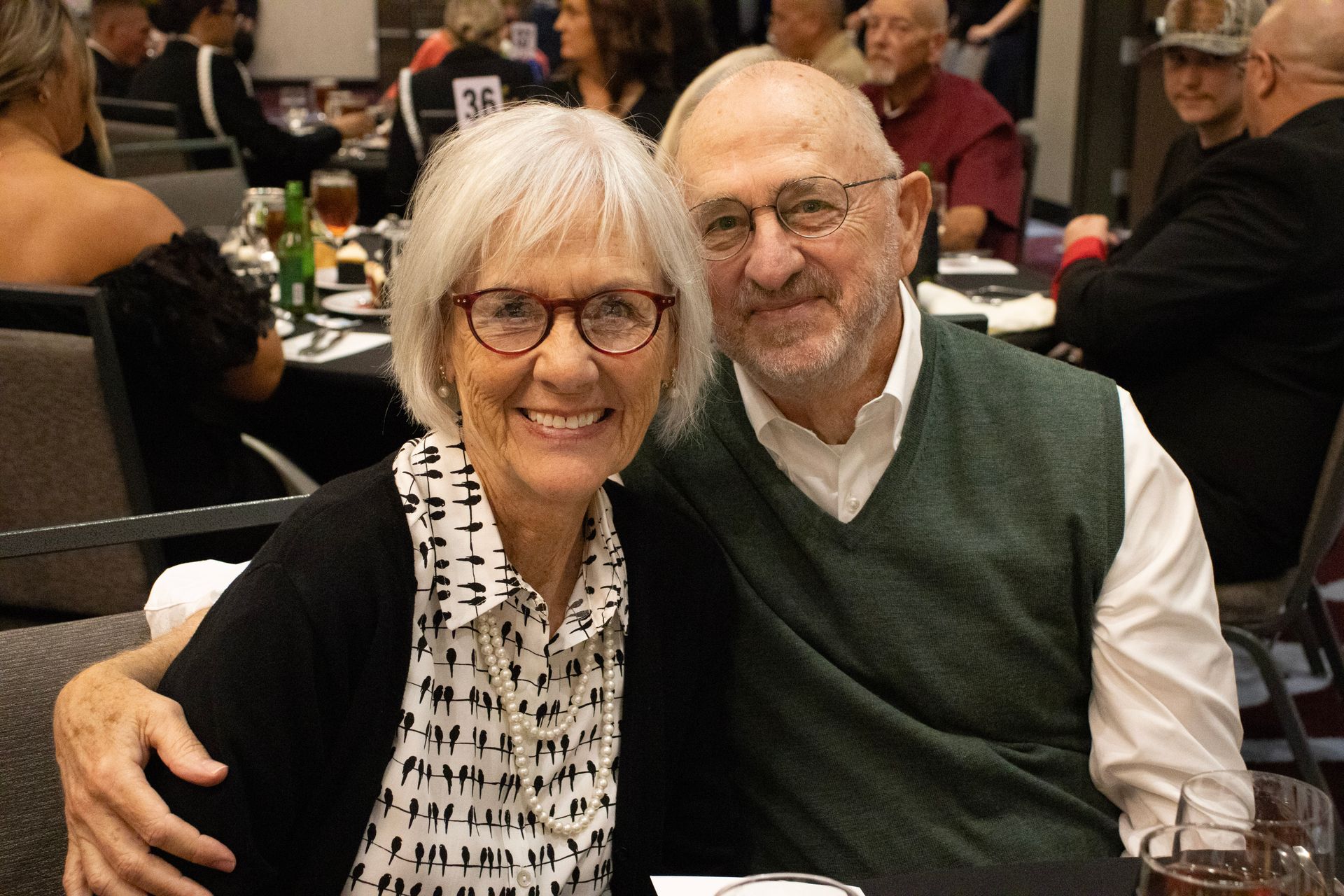 A couple smiling together at a dining table in a banquet hall, both wearing glasses.