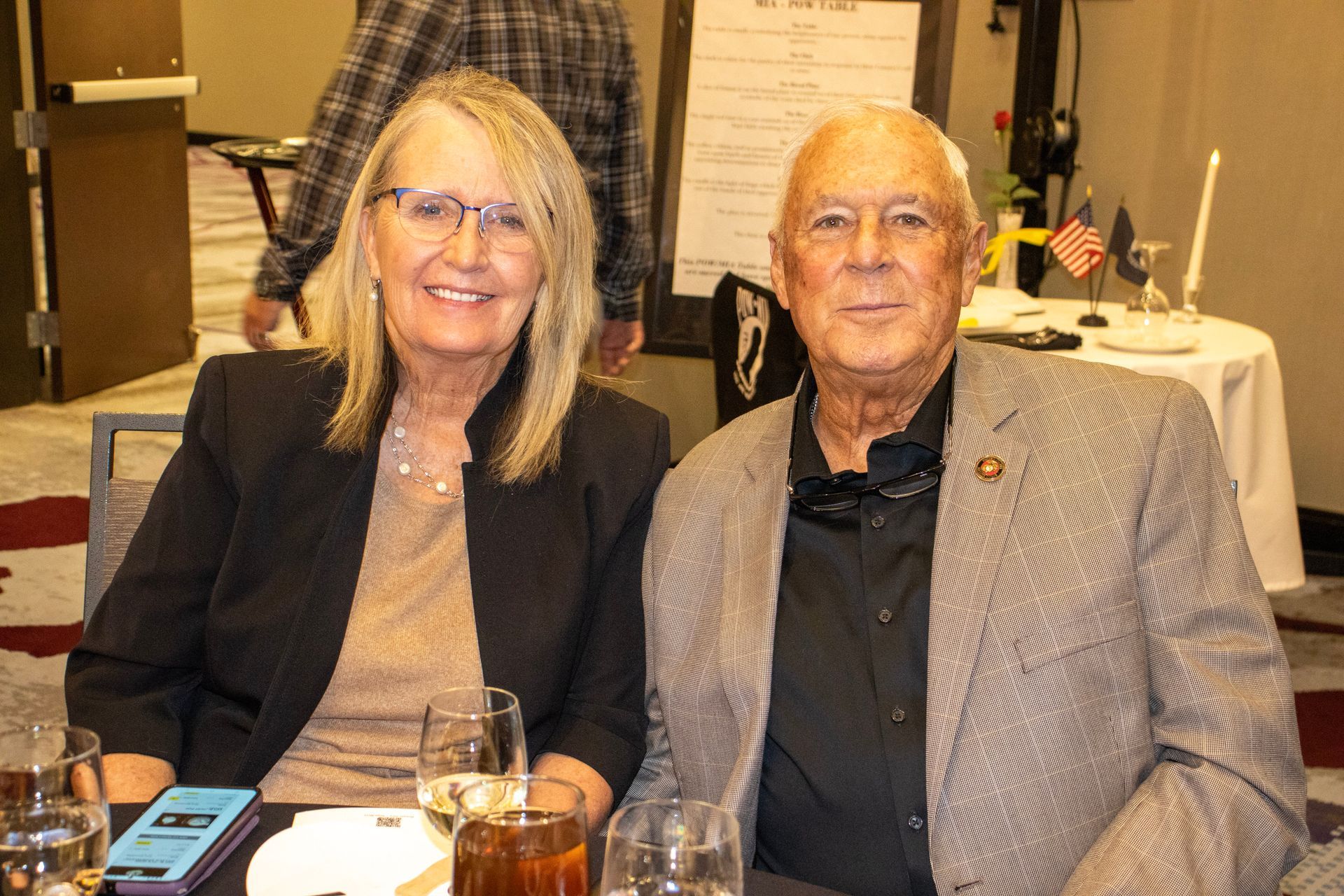 A couple sits side-by-side at a table in a dimly lit room, both smiling at the camera.
