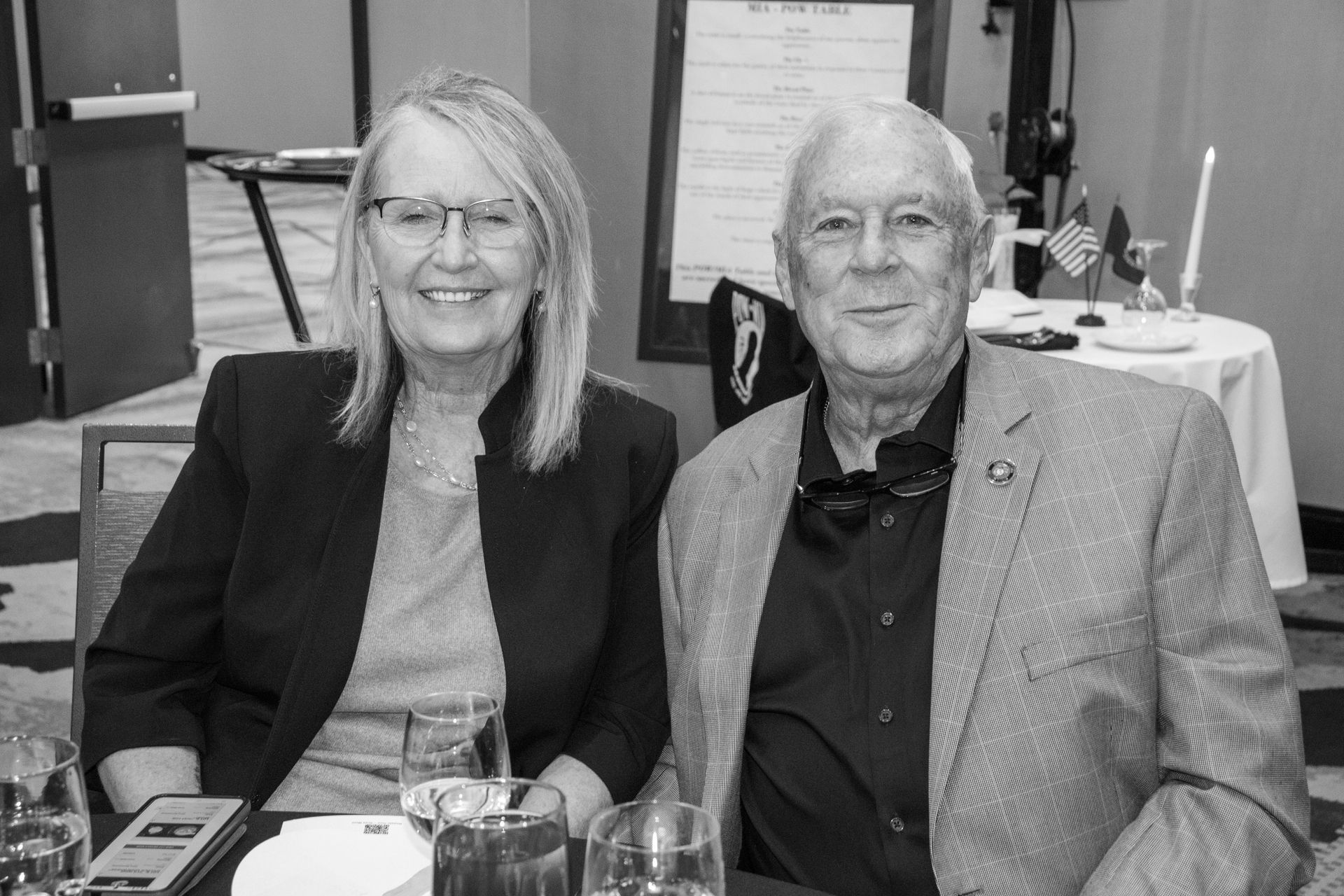 Two people sit at a table in a room with flags in the background, smiling as they pose for a photo.