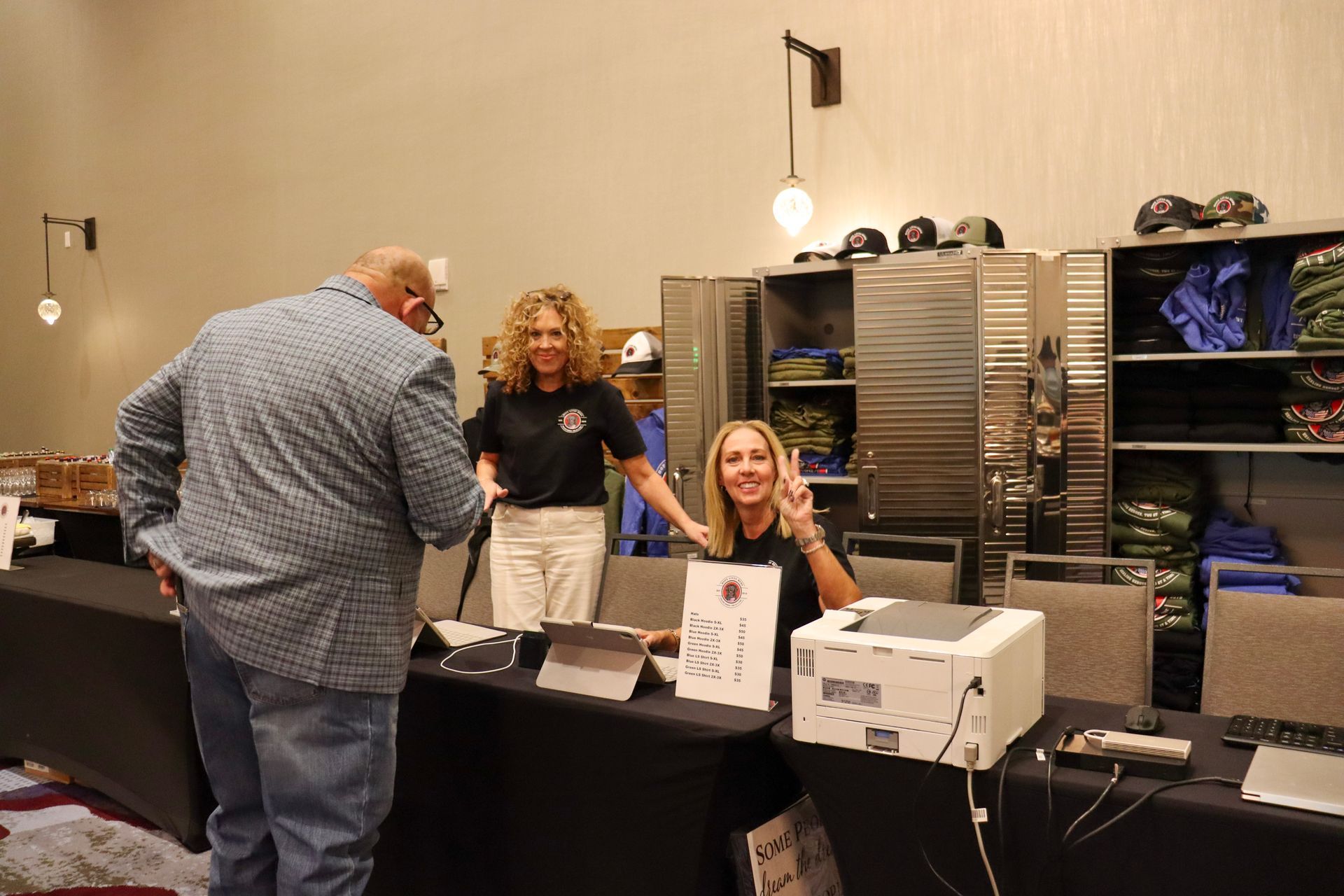 A man in a patterned button-down shirt talks to two people at a registration table with a laptop and printer.
