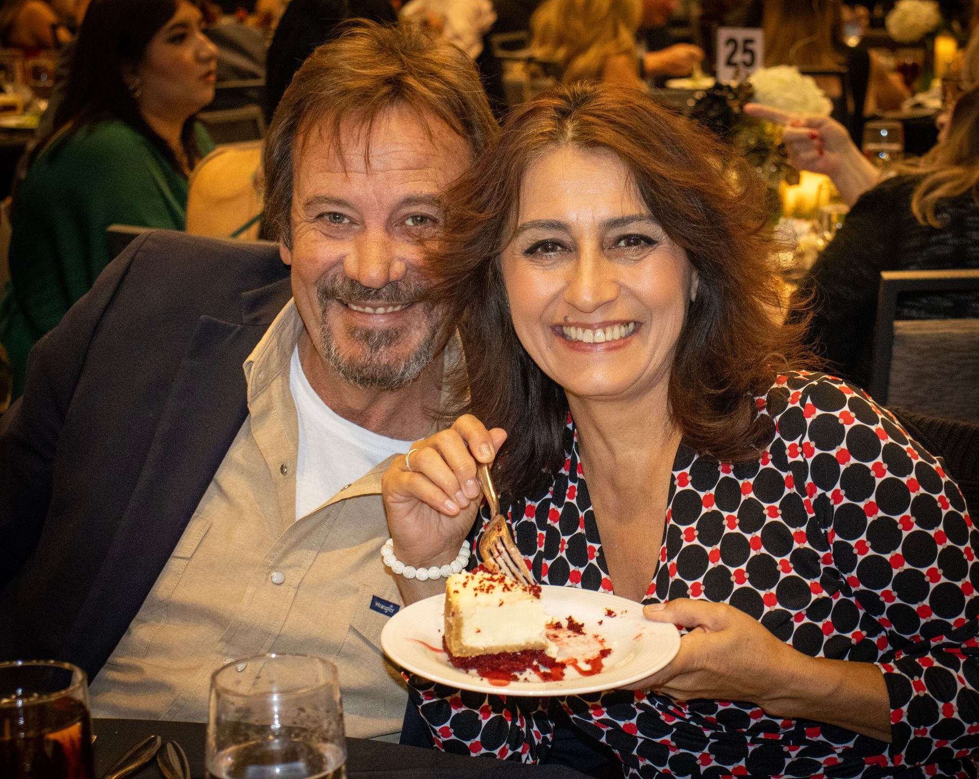 A man and woman sit at a table, smiling while she holds a small plate with a slice of cheesecake at a social event.