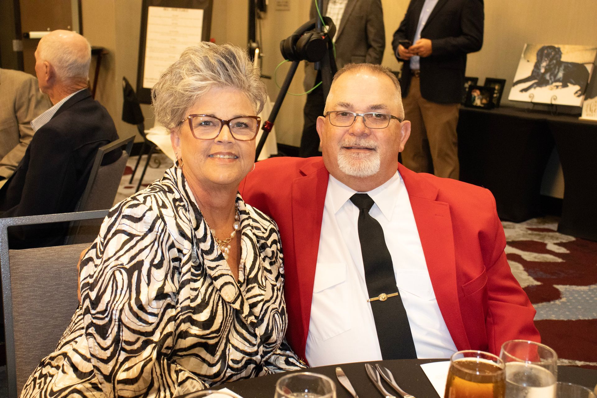 A smiling couple sits together at a table; the person on the left wears a zebra-print top, and the right wears a red blazer.