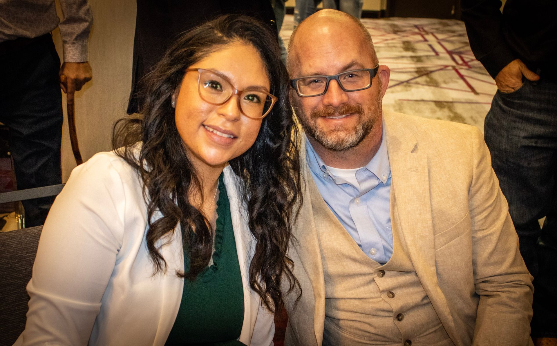 A smiling pair pose together, a woman in a white blazer and green top beside a man in a tan blazer and light blue shirt.