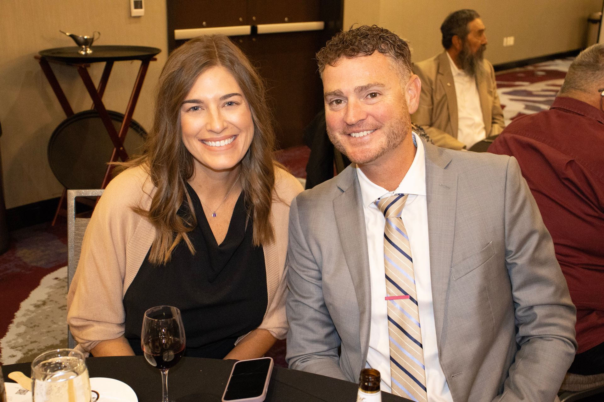 A smiling couple in formal attire pose together at a table during an indoor event.