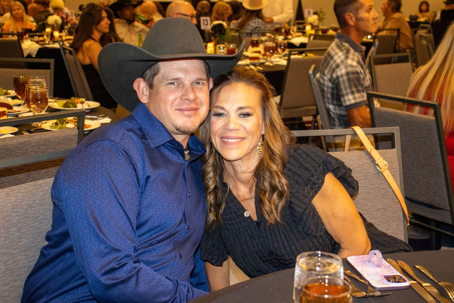 A smiling couple sits together at a dinner table in a banquet hall, with other guests visible in the blurred background.