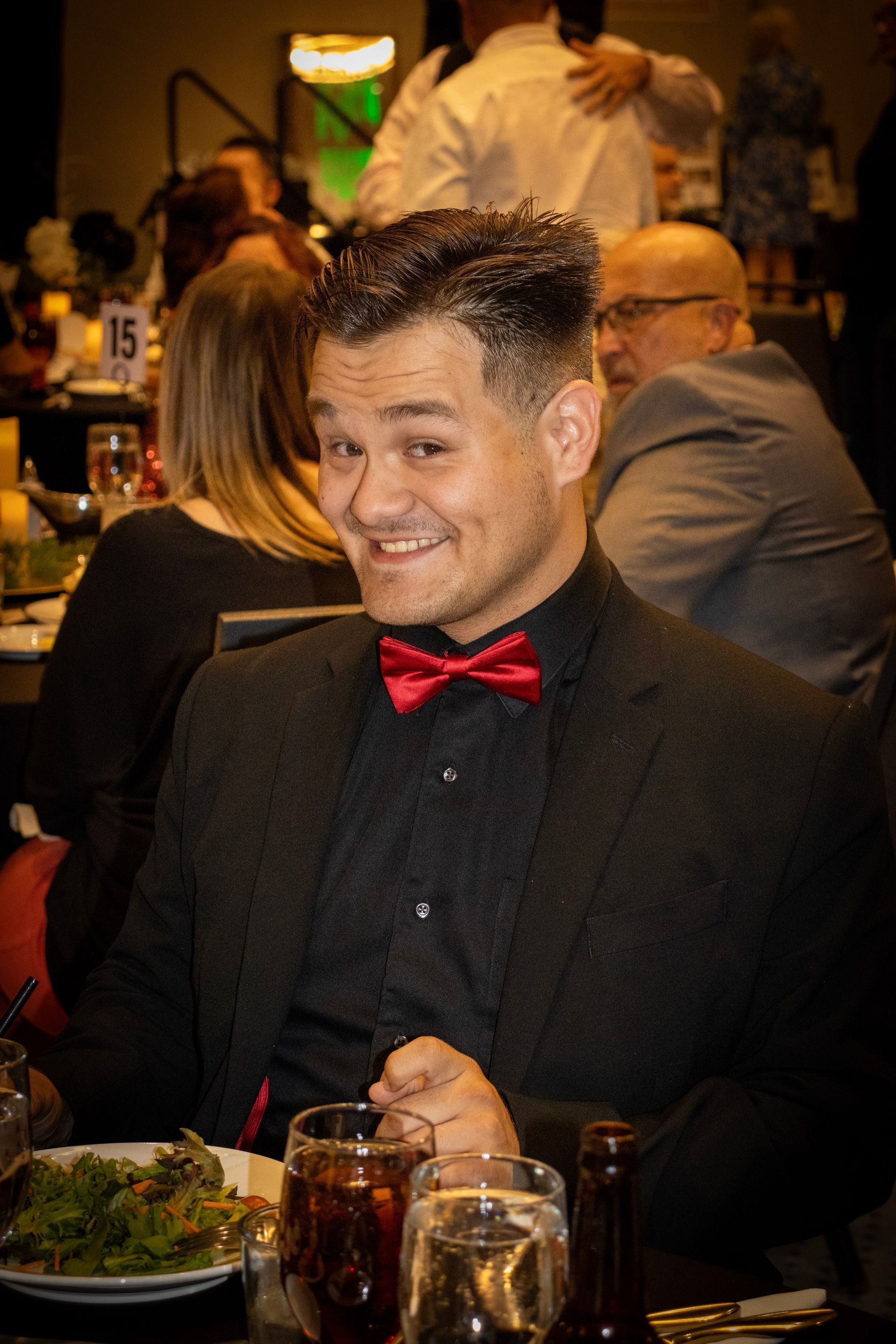 A person with short hair and a red bow tie smiles while sitting at a table in a dimly lit banquet hall.