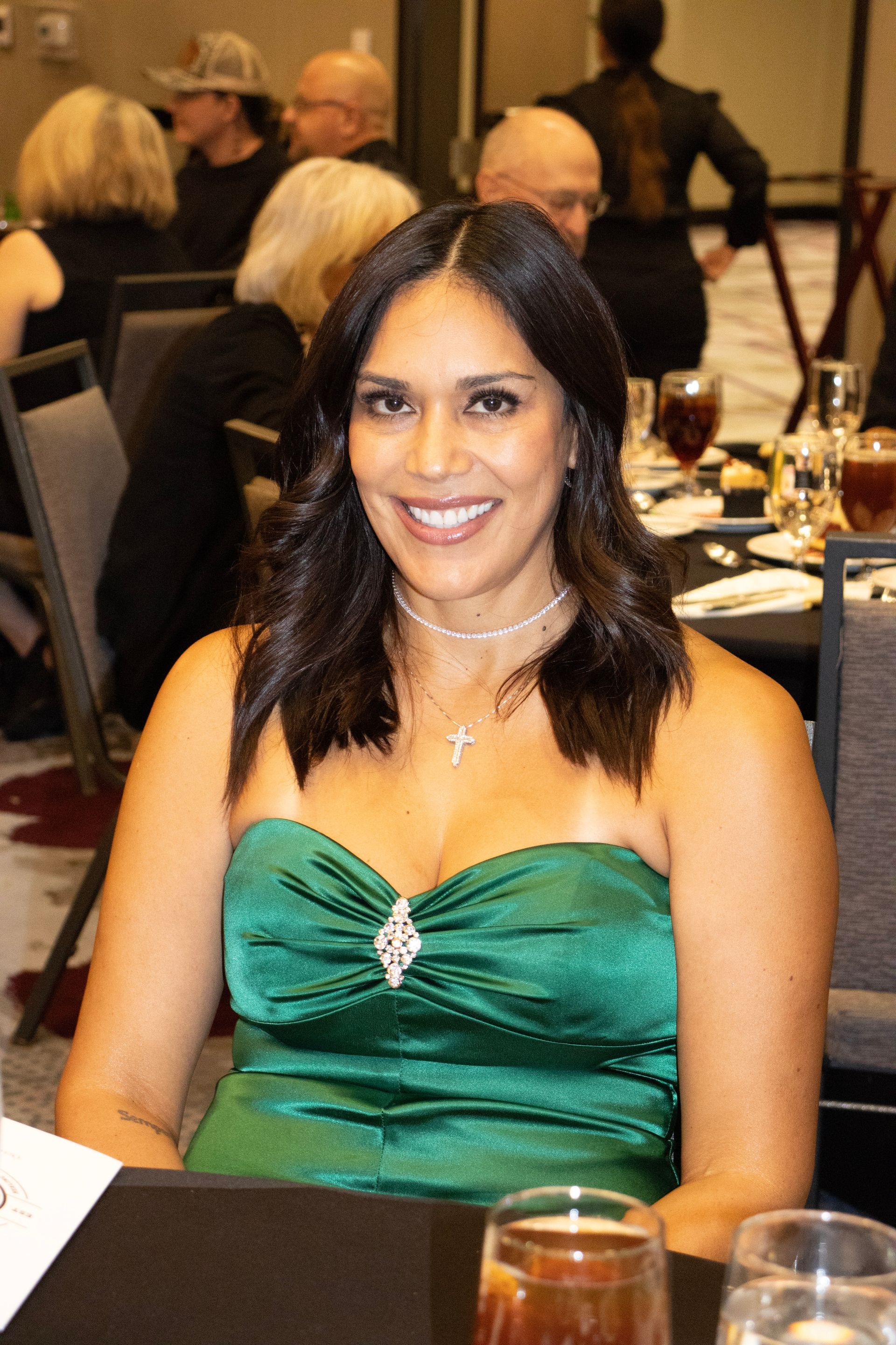 A smiling person wears a green strapless dress and a cross necklace at a banquet table in a dimly lit event space.