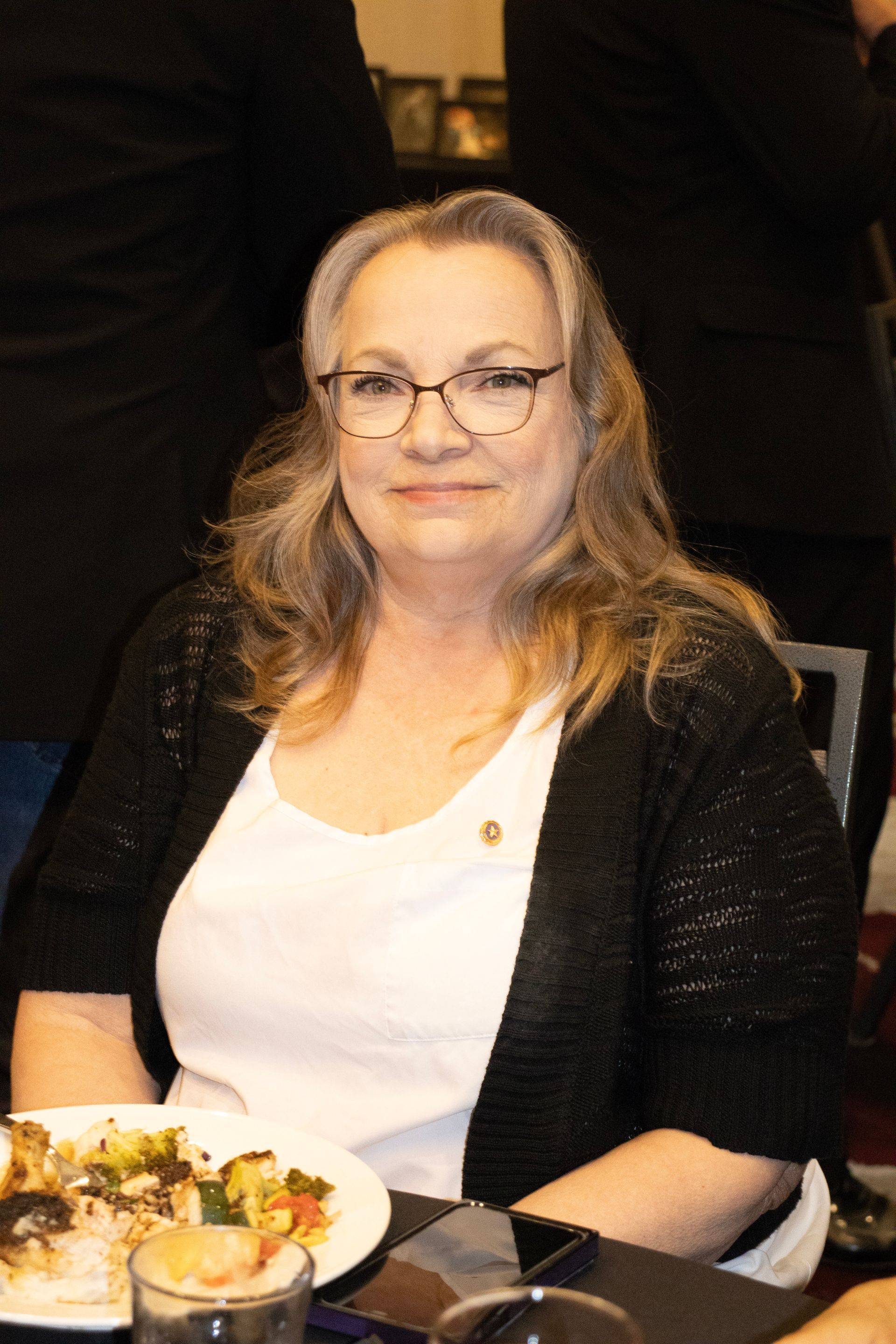 A person with long hair and glasses, wearing a white shirt and black cardigan, sits at a table with food.