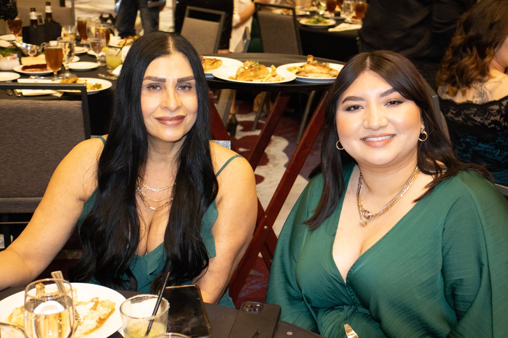 Two smiling women wearing dark green tops pose together at a table in a dimly lit restaurant.