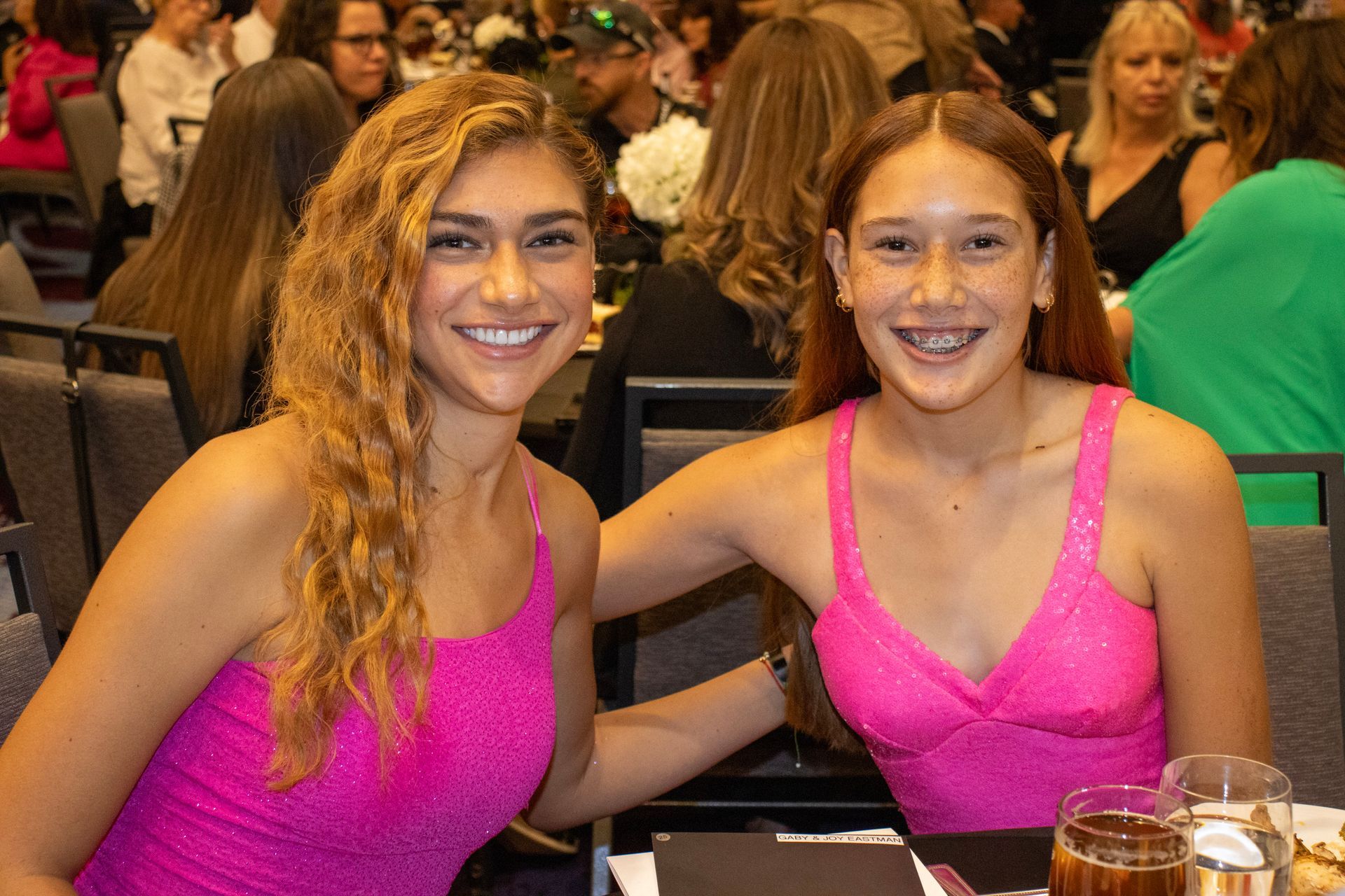 Two smiling people in matching bright pink dresses pose together at a table in a crowded event hall.