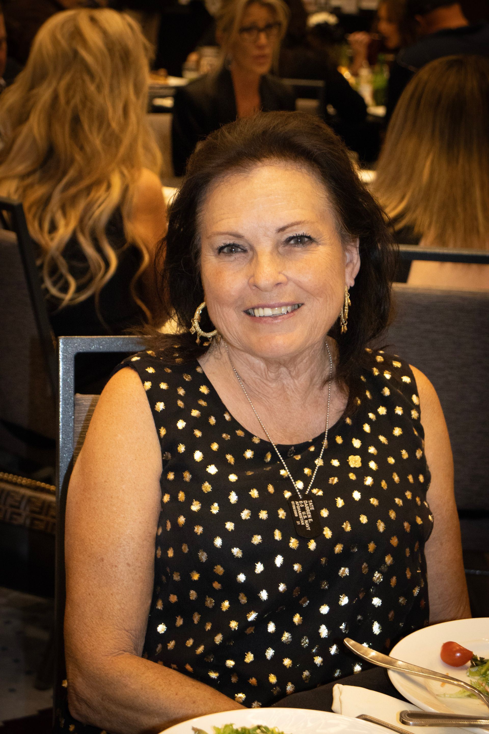 A smiling woman with dark hair wearing a black dress with gold polka dots sits at a table in a dining room.