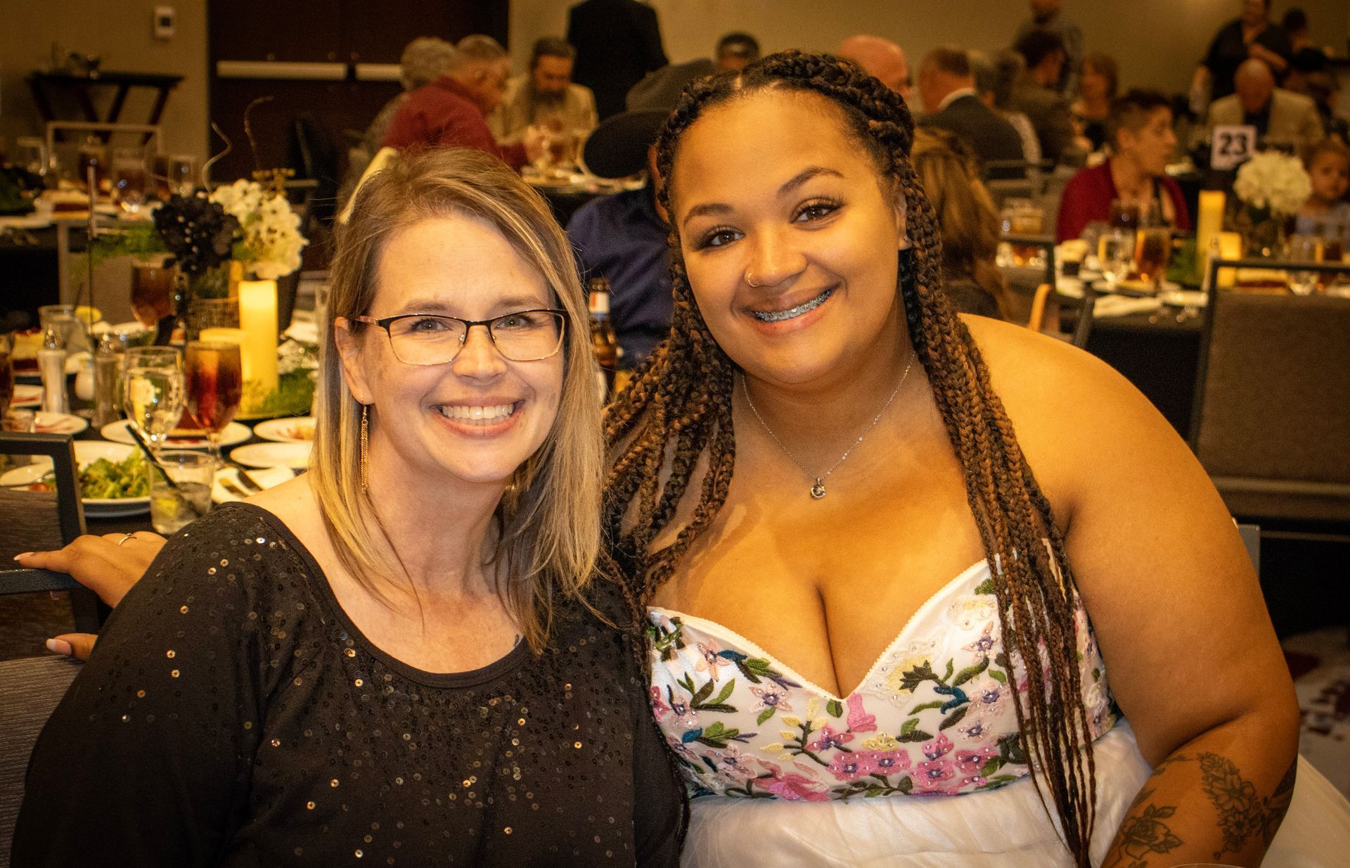 Two women smile while sitting close together at a brightly lit, festive event with tables and guests in the background.
