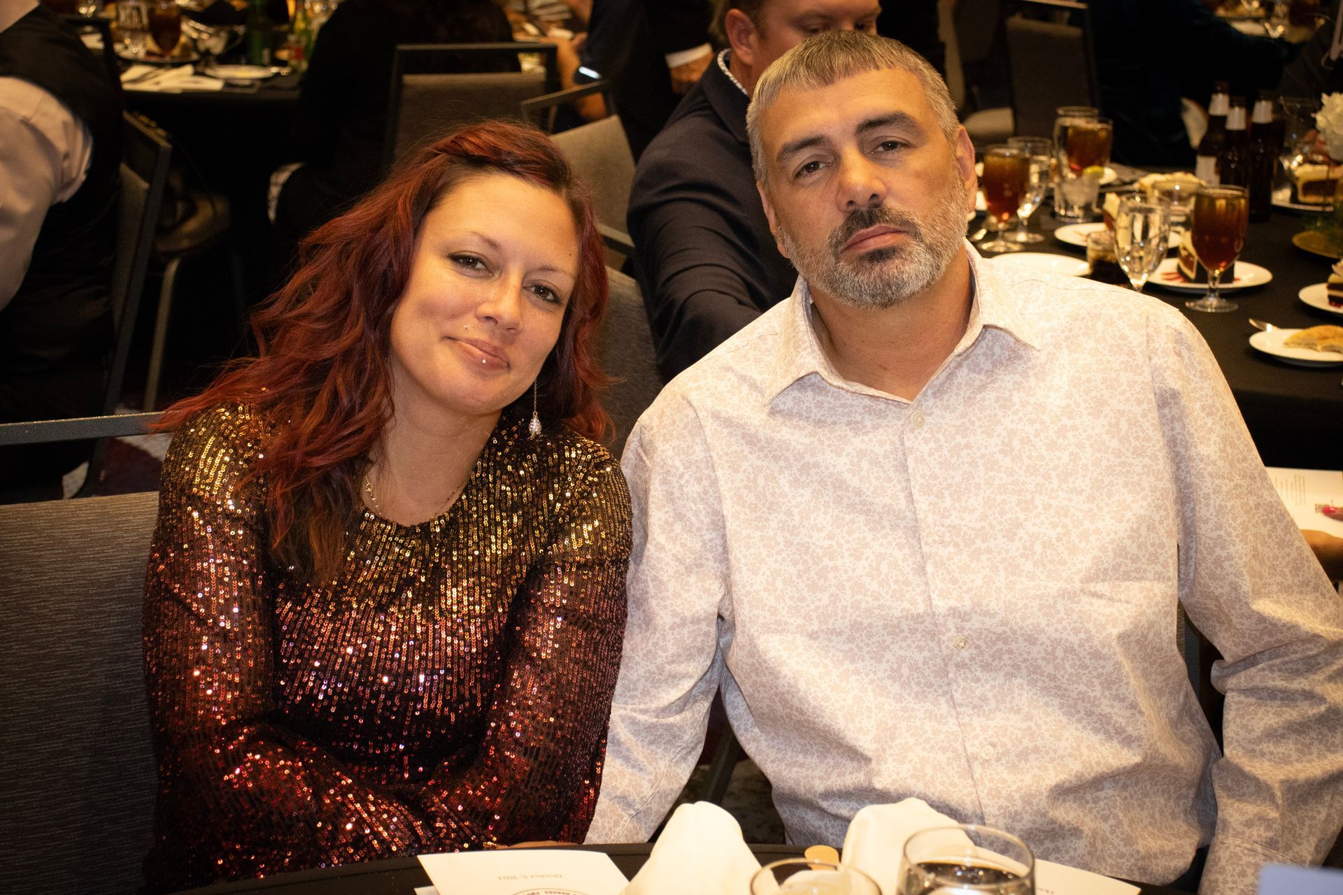 A woman in a shimmering sequined top and a man in a light button-down shirt sit together at a table in a dimly lit venue.