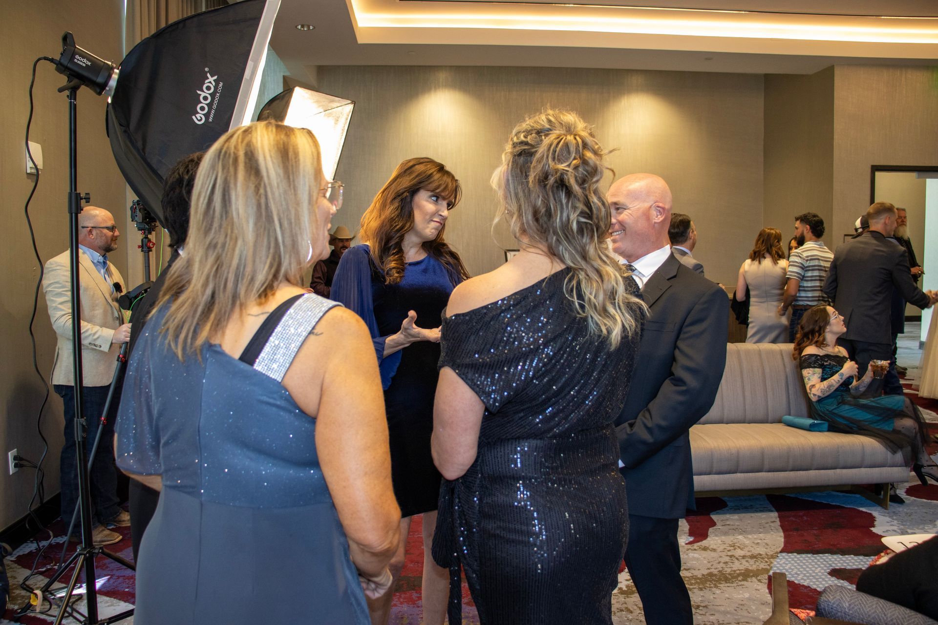 A group of people in formal attire converse in a hotel lobby, with professional photography equipment set up nearby.