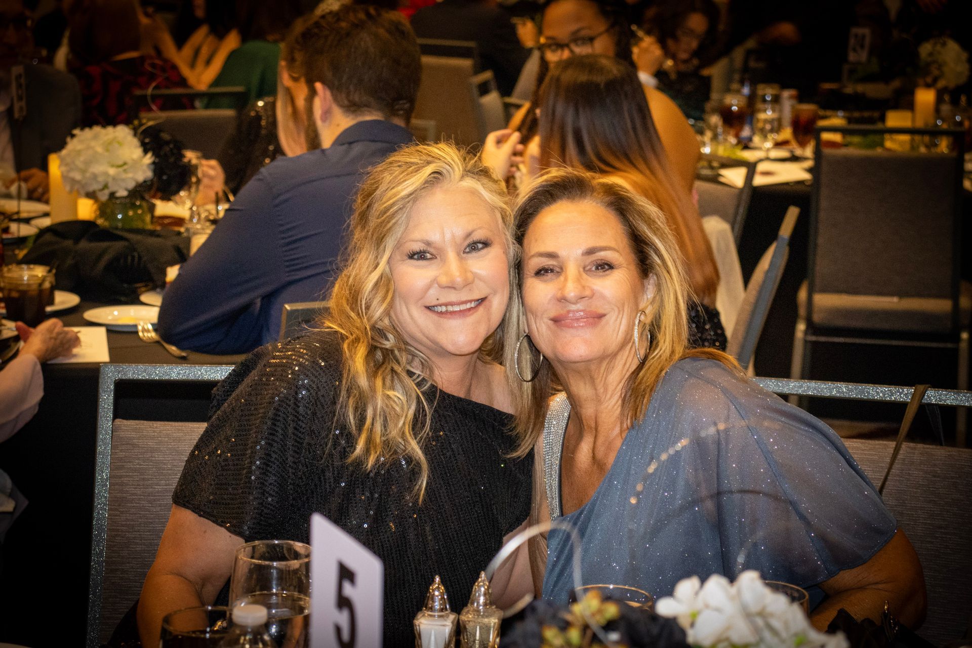 Two women smiling at the camera at a formal dinner event. A table sign with the number five is in the foreground.
