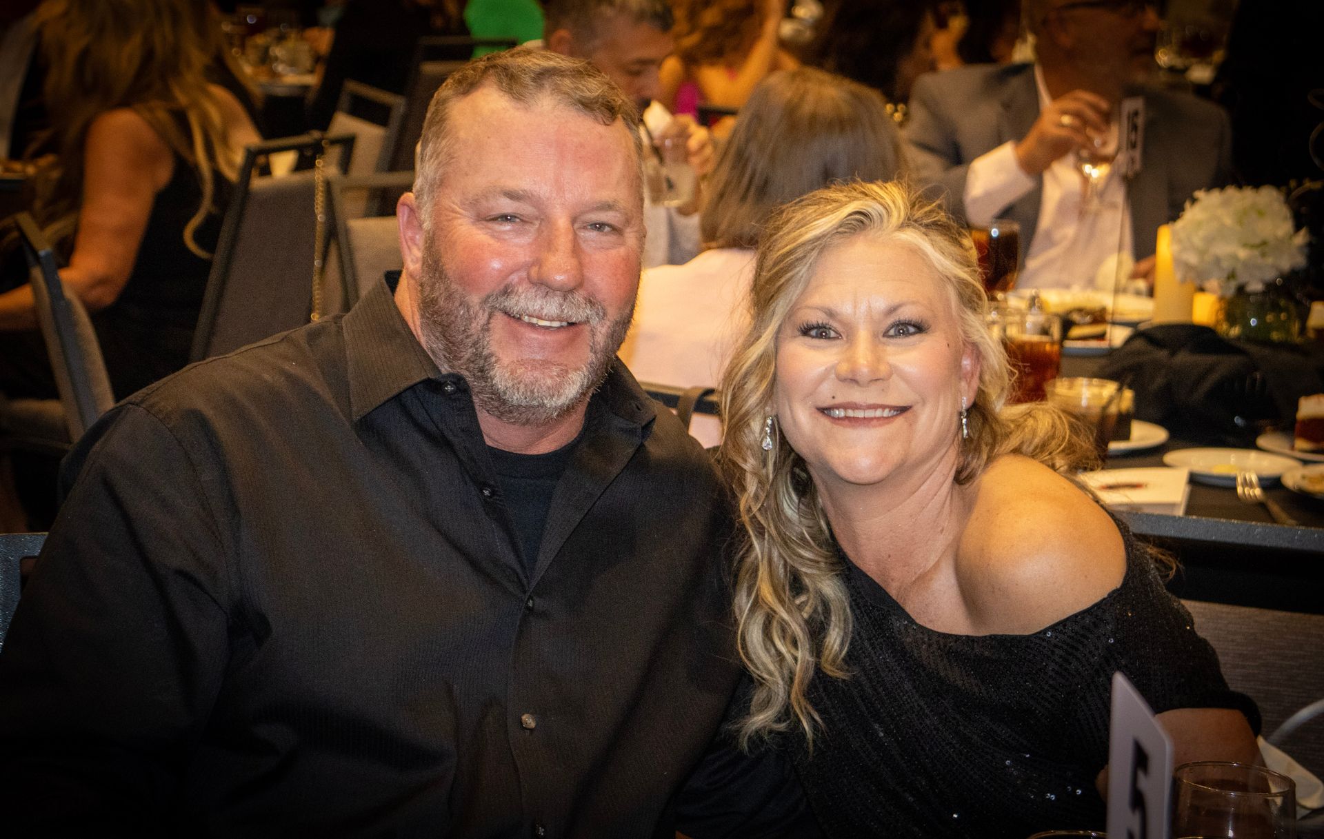 A smiling couple sits together at a dinner event, wearing black formal attire in a crowded banquet room.
