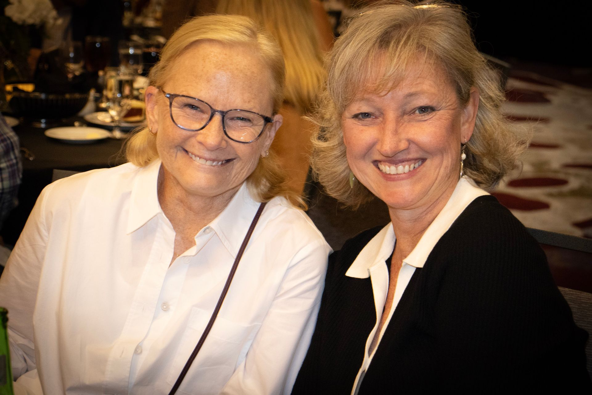 Two smiling women sitting side-by-side at a table in a dimly lit, professional setting.