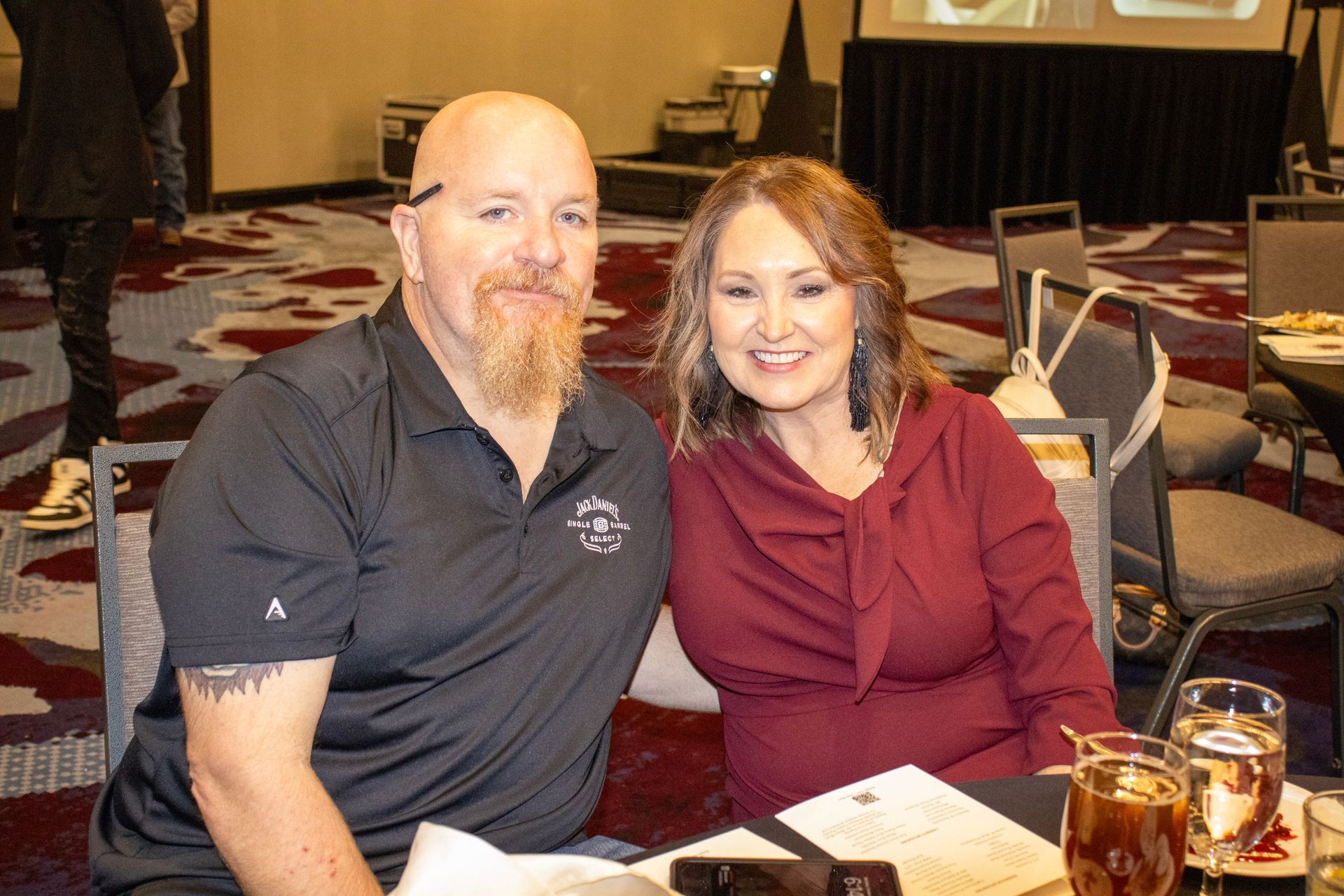 A man in a black polo and a woman in a maroon blouse smile while sitting at a table in a conference room.