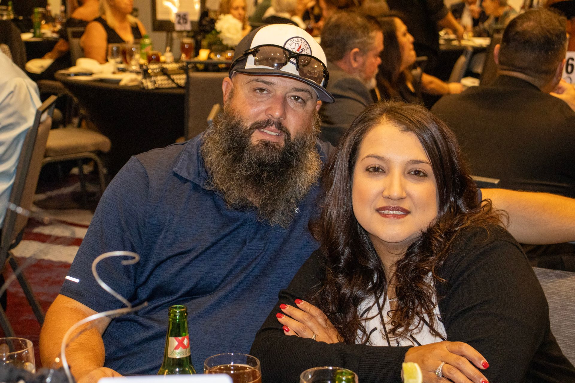 A person with a beard and baseball cap sits beside a person with dark hair at a table in a dimly lit, social setting.